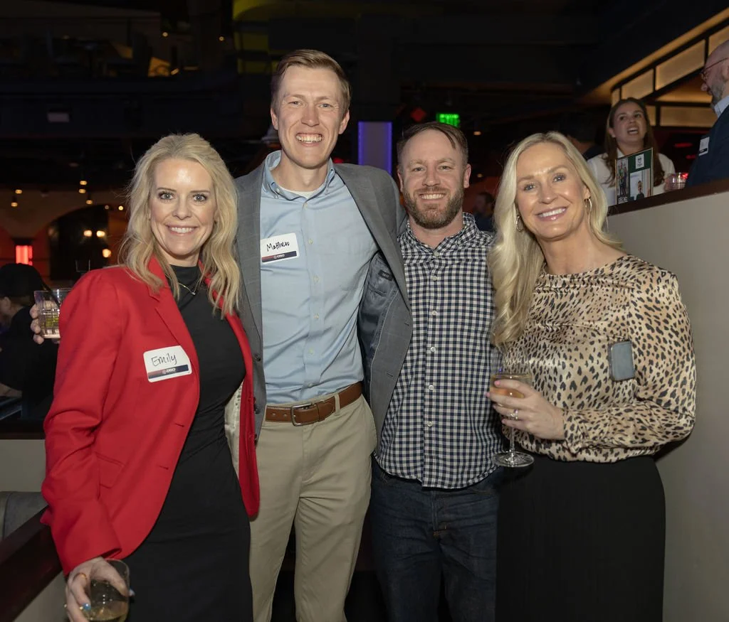 Four people smiling and posing together at a social event, holding drinks, in a dimly lit venue with a bar in the background. A woman in a red blazer, a tall man in a gray blazer, a man in a checkered shirt, and a woman in a leopard-print blouse.