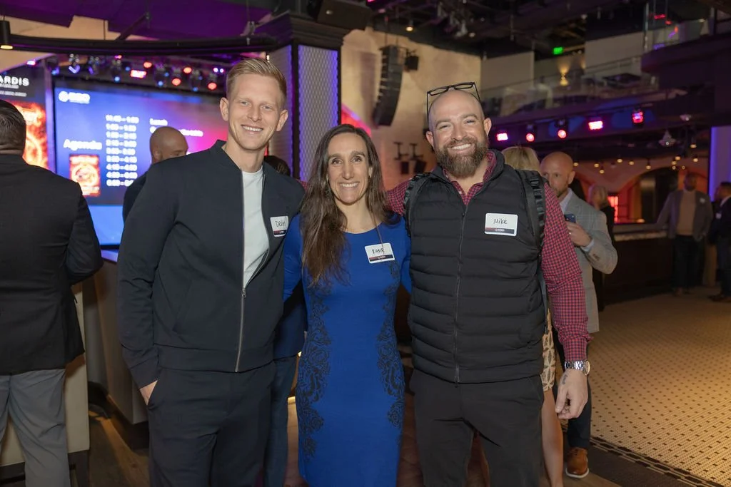 Three smiling people posing together at an indoor event, with a bar and a large screen displaying a schedule or menu in the background.