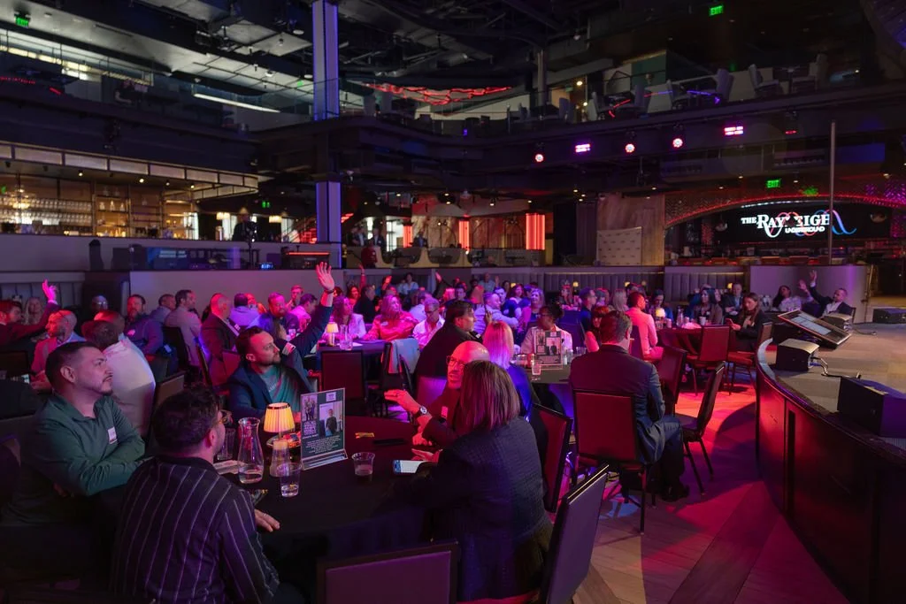 A large crowd of people seated at tables in a dimly lit nightclub or comedy club, with a stage on the right and neon signs in the background.