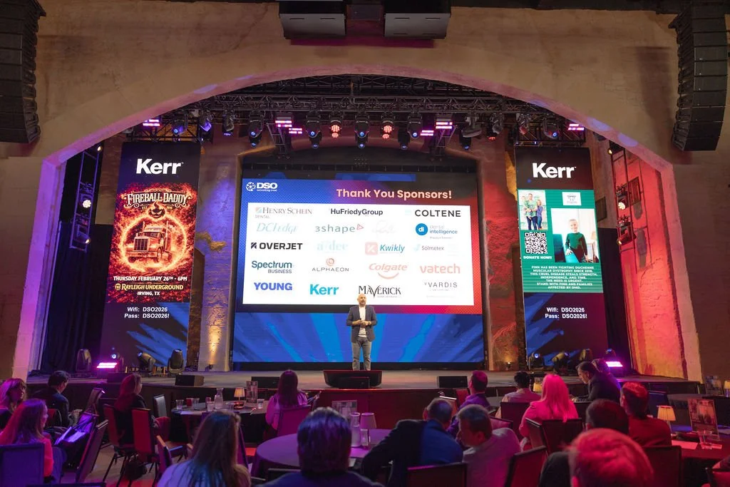 A speaker on stage at a conference or awards event with a large screen behind displaying numerous sponsor logos and a thank you message. The event has a dark, professional atmosphere with an audience seated in front.