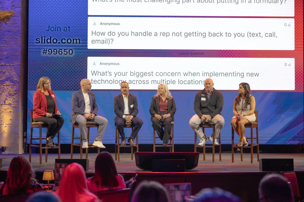 A panel of five people seated on a stage with a large screen behind them displaying questions from an online audience. The stage has a textured stone wall on the left, and the audience is visible in the foreground.
