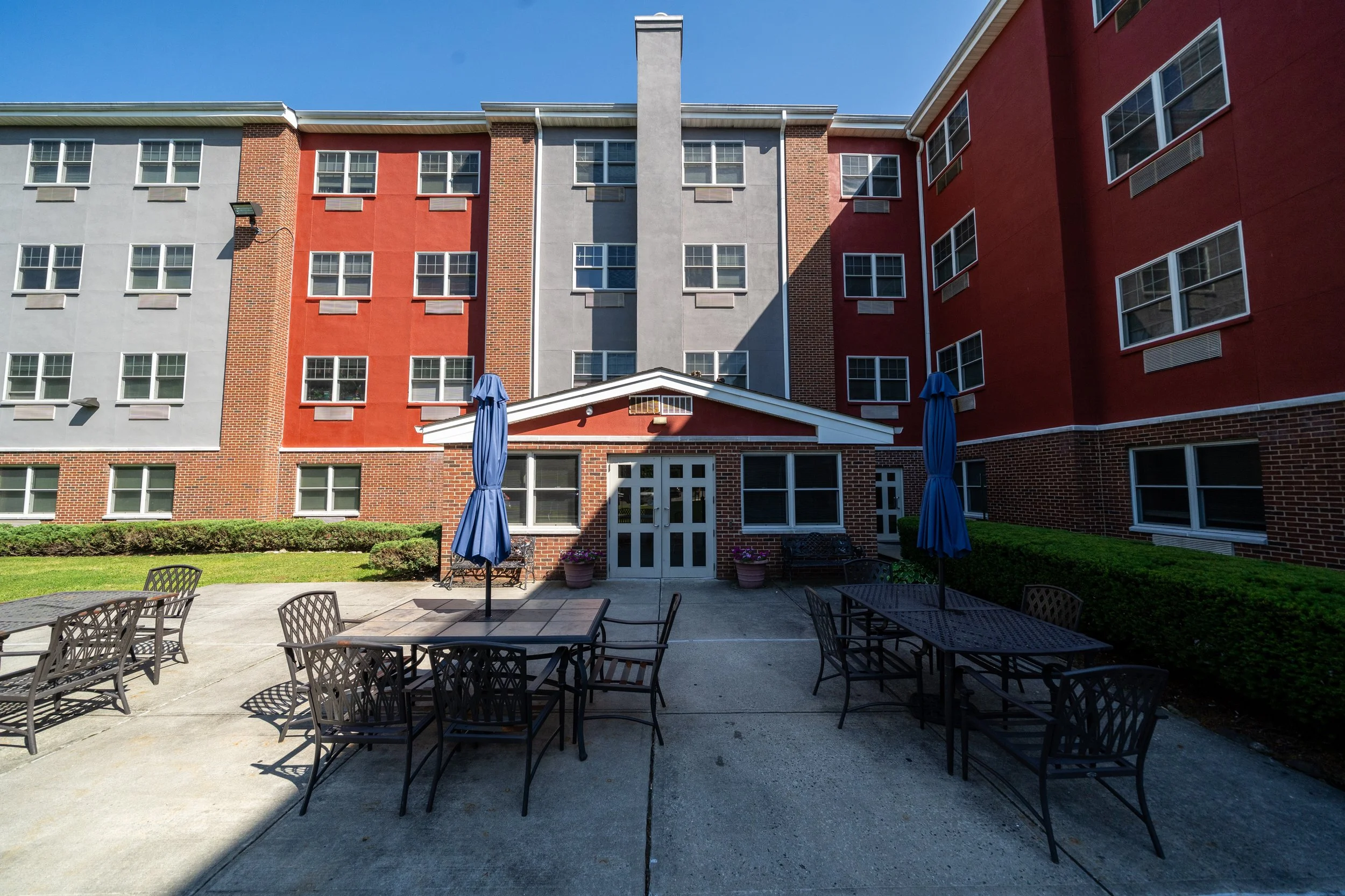 Outdoor patio area with metal tables, chairs, and closed blue umbrellas in front of a multi-story apartment building with red, gray, and brick exterior