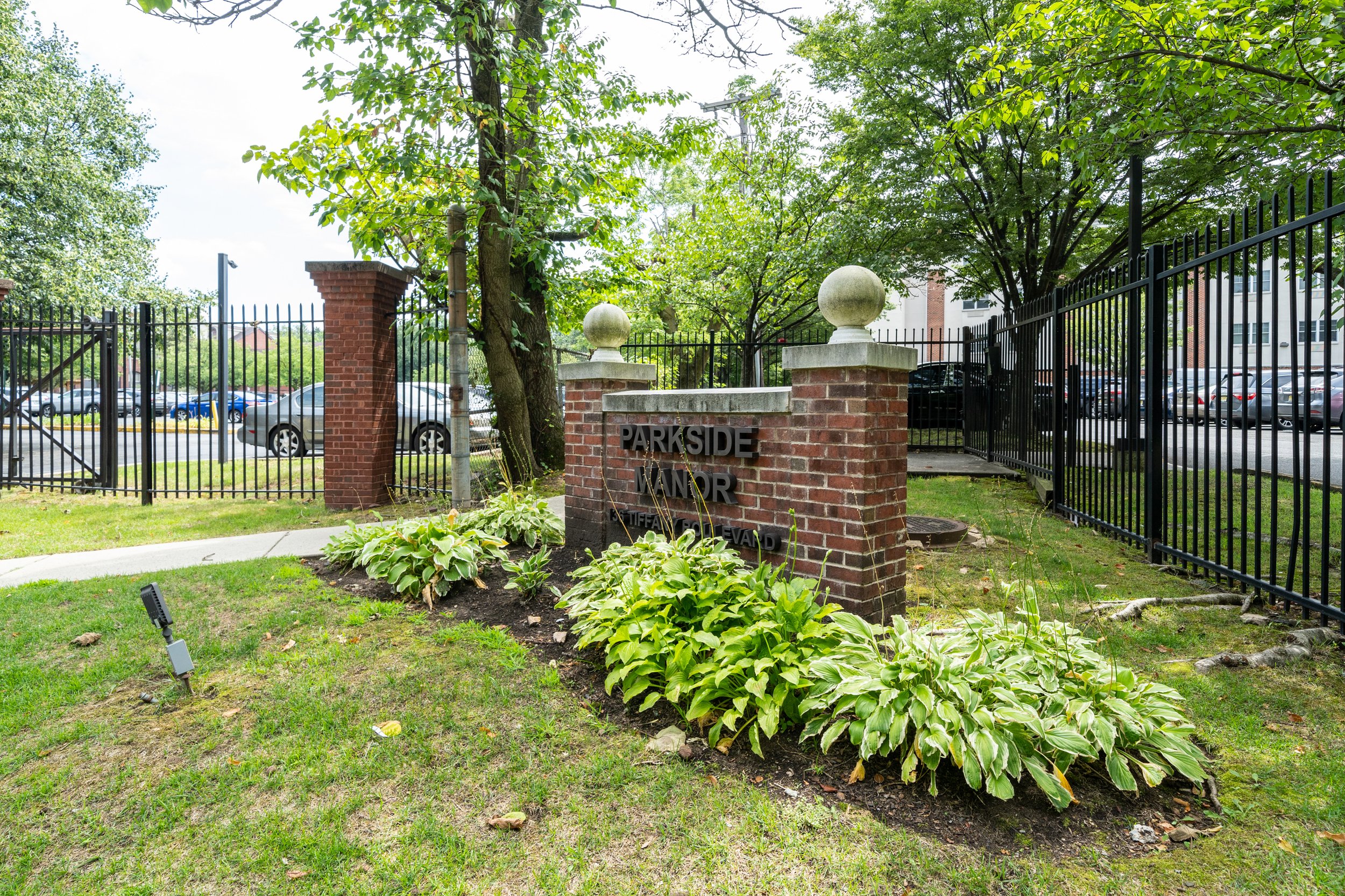 Brick entrance sign reading 'Parkside Manor' surrounded by greenery and a parking lot with cars.
