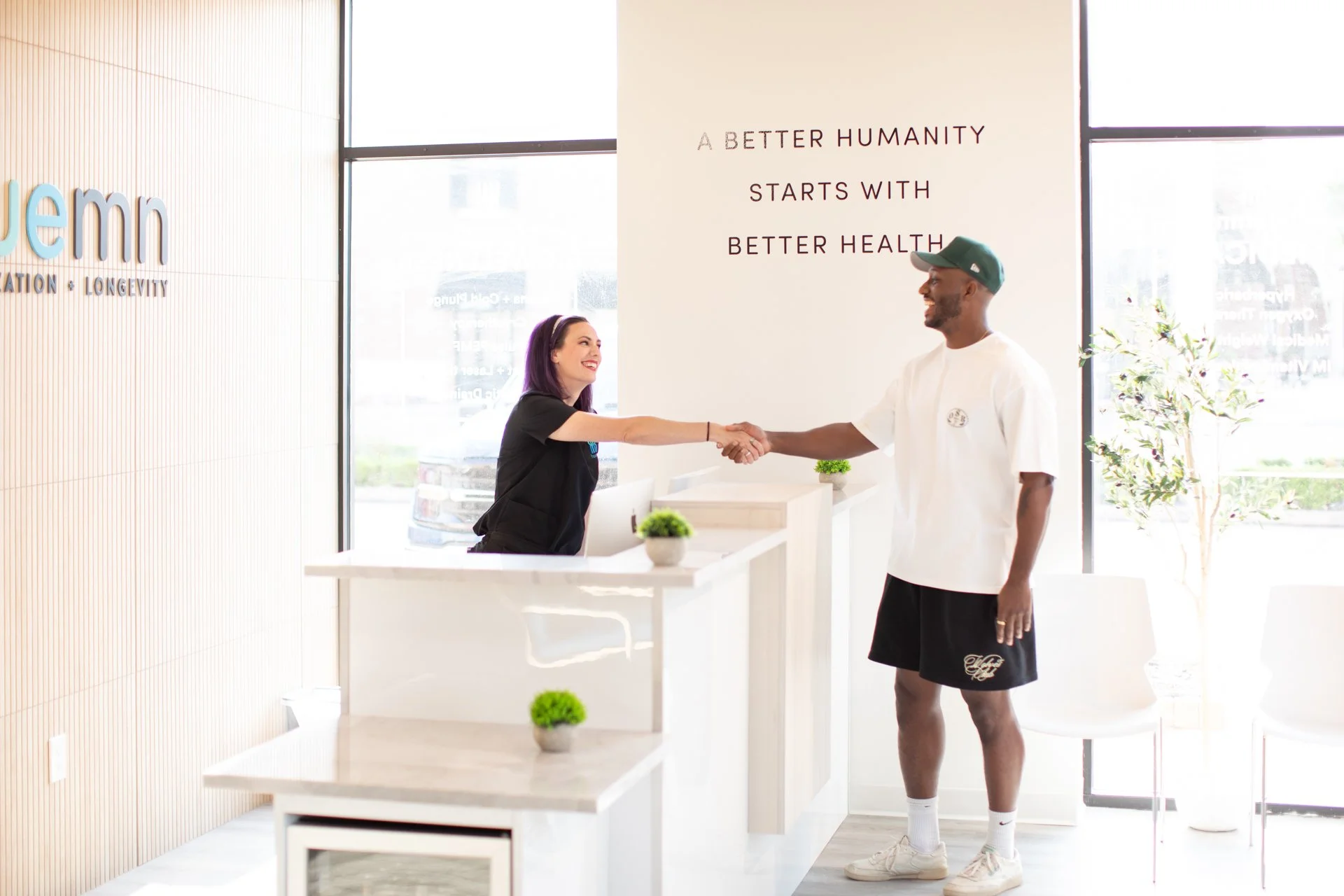 A woman and a man shaking hands at a reception desk inside a wellness or healthcare center. The woman has purple hair and is smiling. The man is wearing a white t-shirt, black shorts, white sneakers, and a green cap. Behind them, a sign on the wall reads, "A BETTER HUMANY STARTS WITH BETTER HEALTH." The reception area is minimalistic with small plants and large windows letting in natural light.