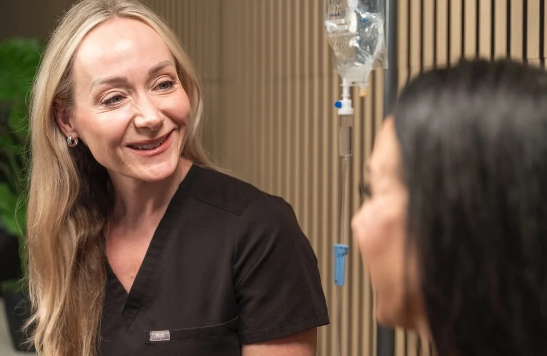 A smiling woman in black scrubs talking to a woman with dark hair in a medical setting with an IV drip in the background.
