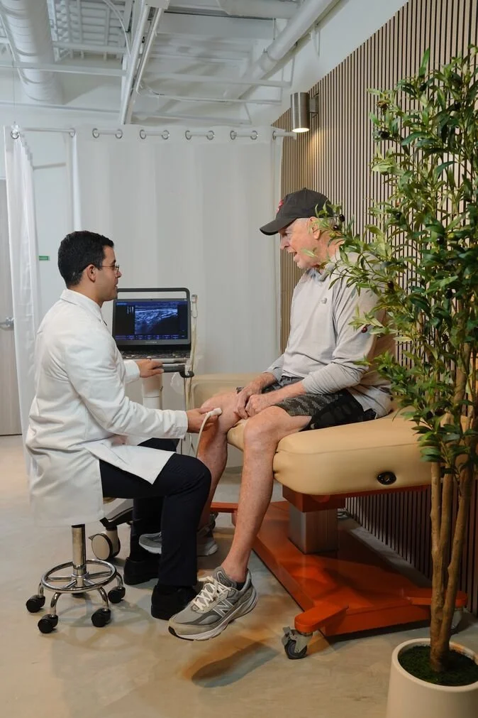 A doctor in a white coat performs an ultrasound on the knee of an older male patient wearing a baseball cap, within a clinical setting beside text explaining "Stem Cell Therapy at Huemn."