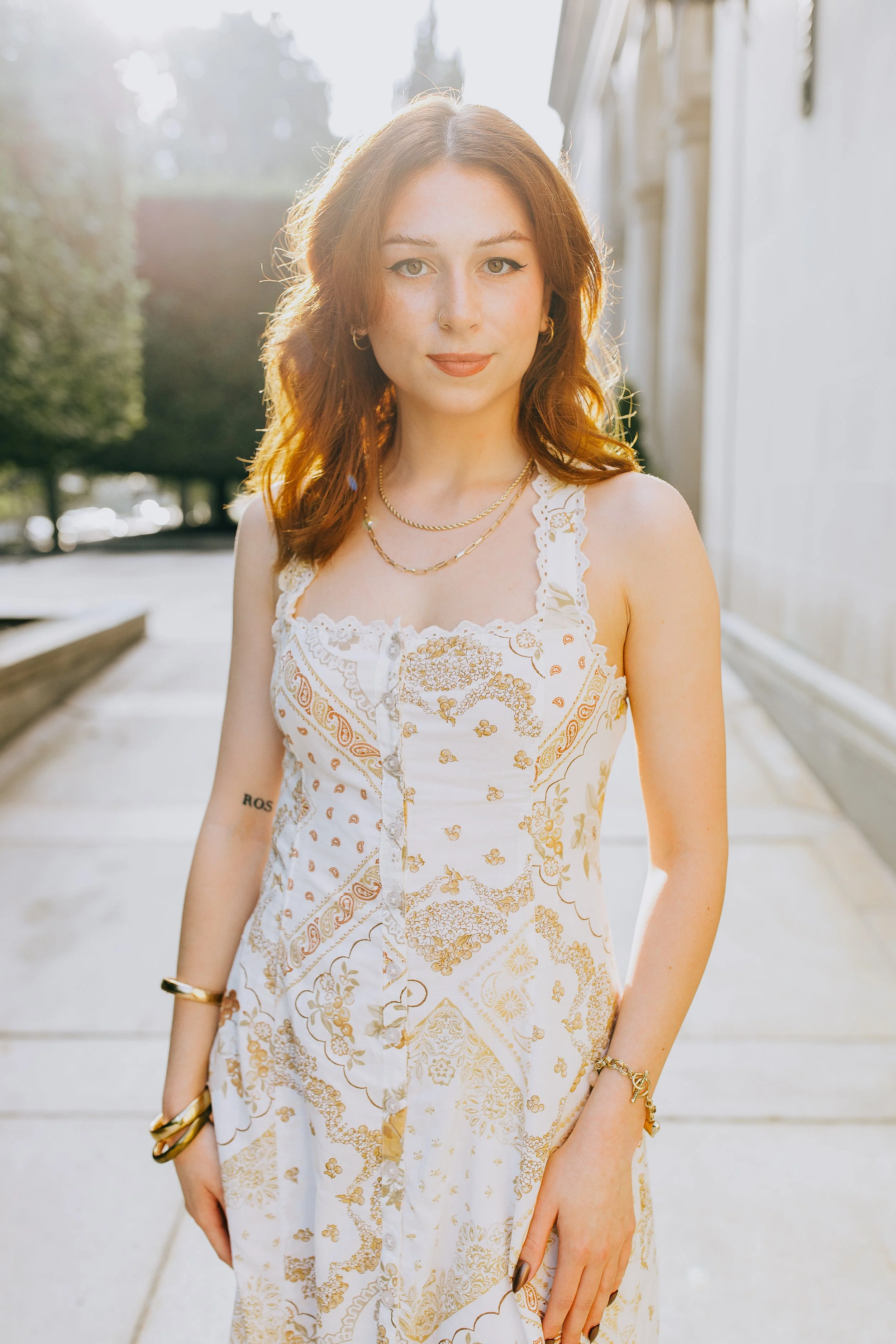A young woman with red hair and light skin standing outdoors on a sunny day, wearing a white and gold patterned sleeveless dress, gold jewelry, and a slight smile.