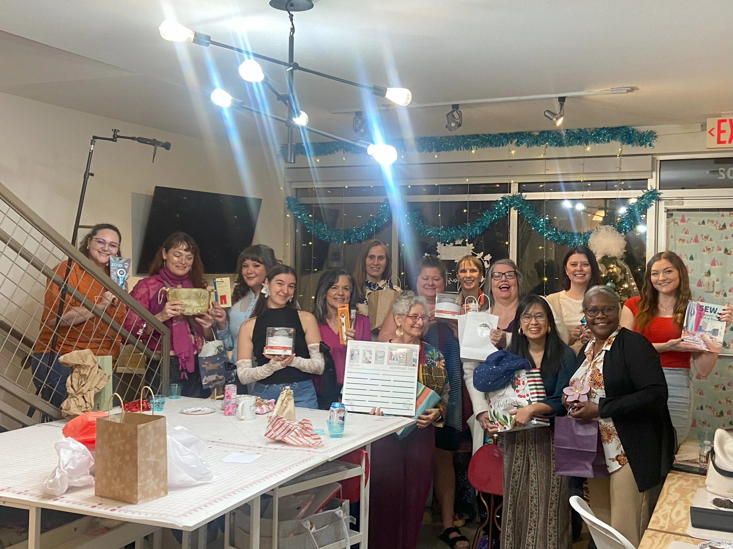 A group of women gathered at a Christmas celebration, smiling and holding gifts. The room is decorated with blue tinsel and Christmas ornaments, with a large window and an exit sign in the background.