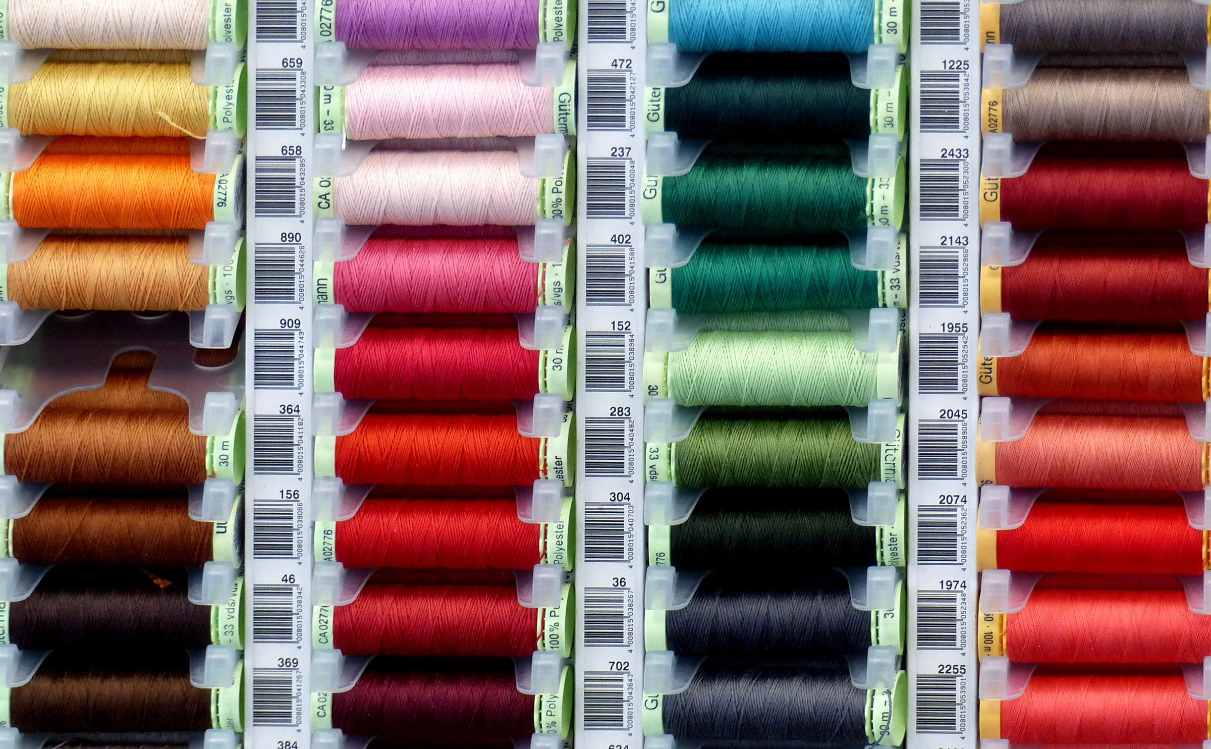 Rows of colorful sewing thread spools organized by color, including yellow, pink, red, green, blue, gray, black, and orange, with barcode labels in the forefront.