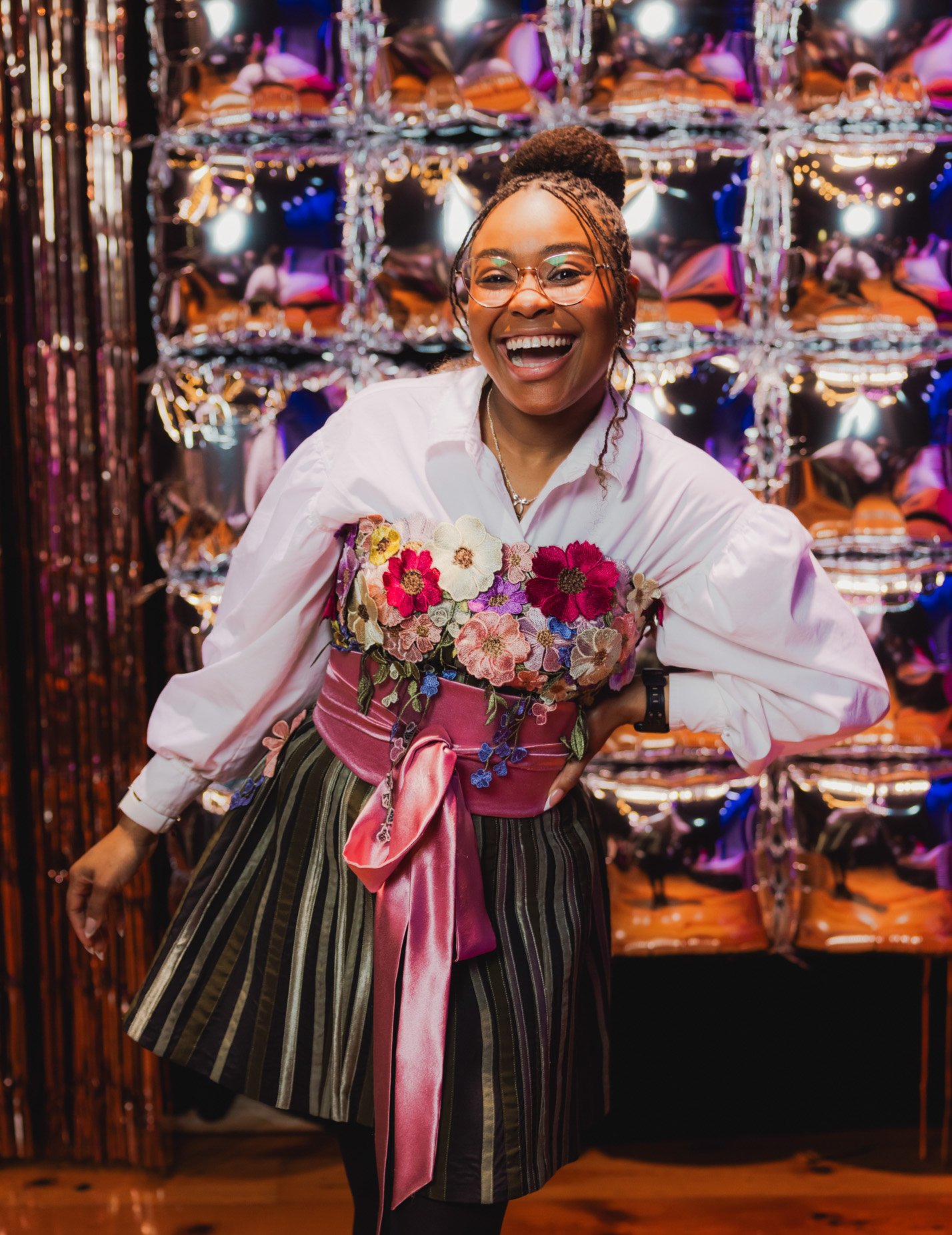 A woman with glasses and braids, smiling, wearing a white blouse and a colorful embroidered corset with flowers, in front of a shiny, metallic, balloon-like backdrop at a festive event.