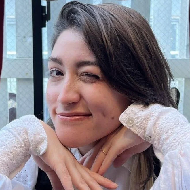 A young woman with dark brown hair, winking and smiling with one cheek slightly raised, resting her chin on her hands while wearing white lace gloves and a white top, in a setting with large window panes behind her.