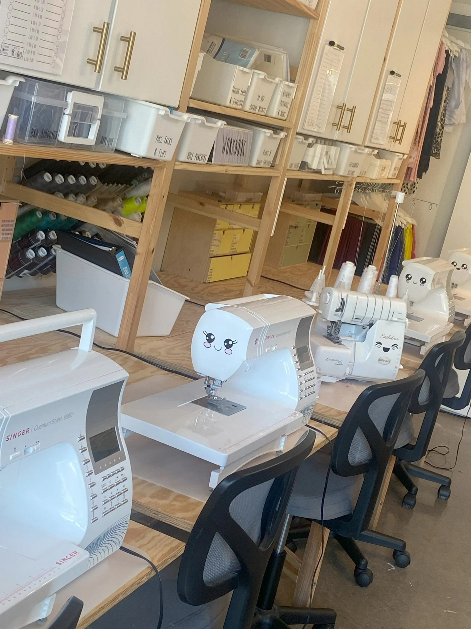 A sewing workshop with multiple computerized sewing machines, some with smiling face covers, in front of wooden shelving units filled with fabric, thread, and labeled storage boxes.