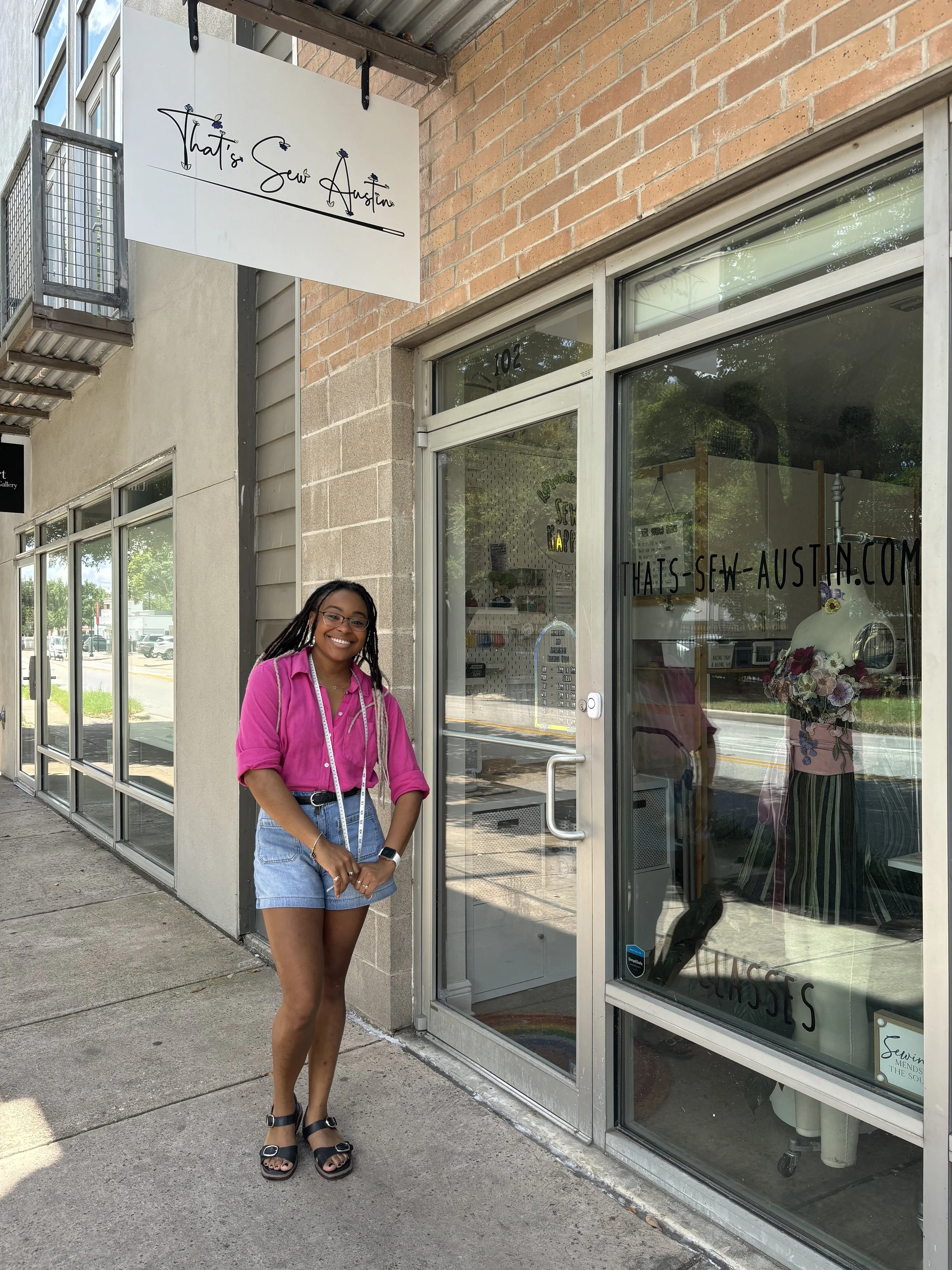 A woman in a pink blouse and denim shorts standing outside a sewing shop named 'That's Sew Austin.' The shop has a glass door with the shop's website written on it and a window display with sewing equipment and a mannequin with flowers.