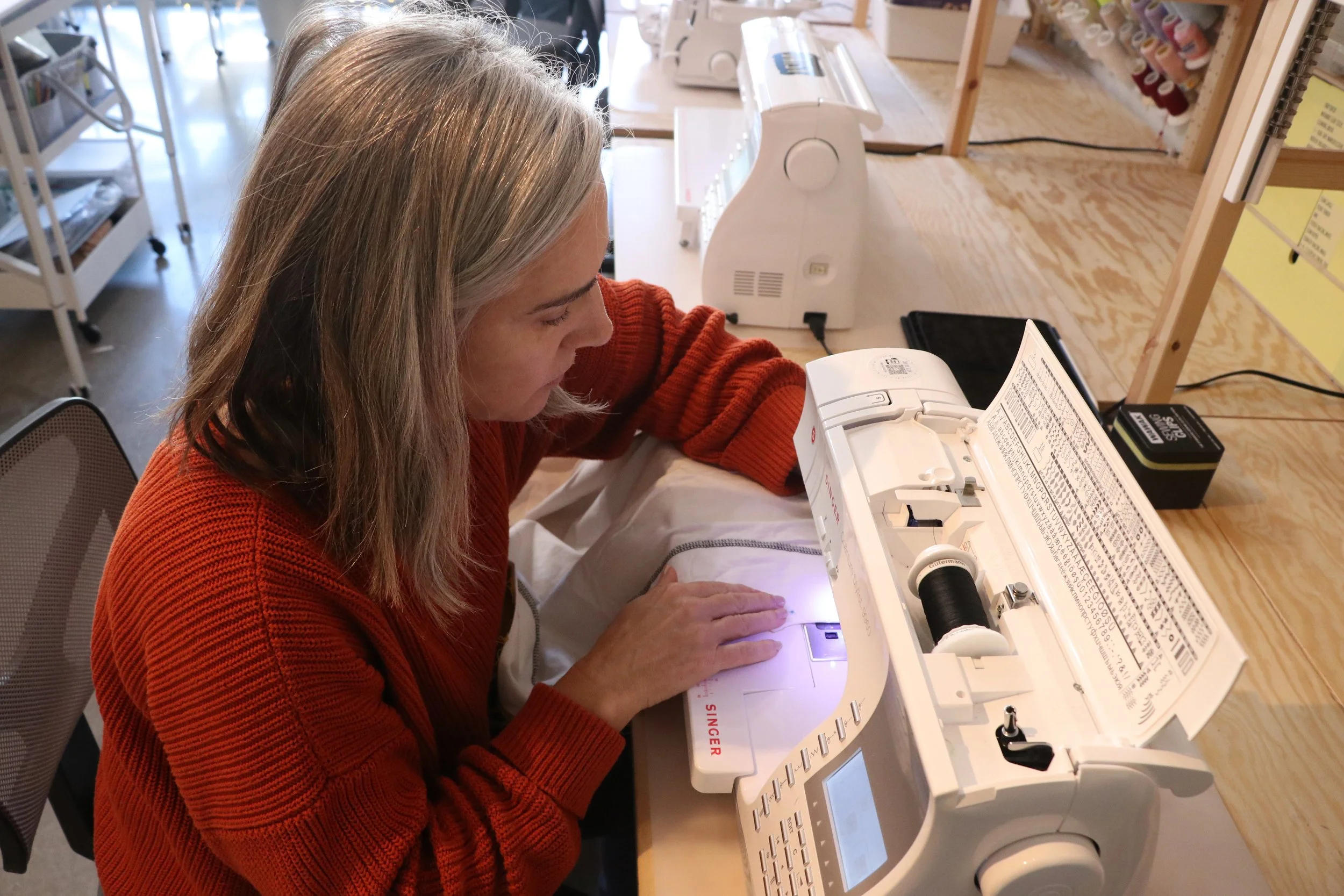 A woman using a sewing machine at a wooden worktable, surrounded by sewing supplies and machines in a craft room.
