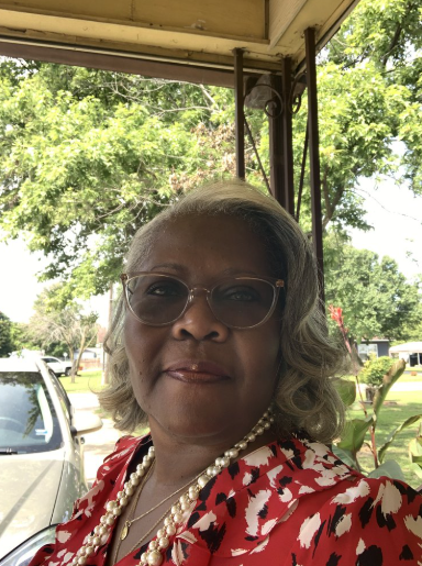 A woman with gray hair wearing sunglasses, a red and white patterned top, and a pearl necklace, standing outside on a porch with green trees in the background.
