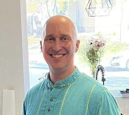 A smiling man with a shaved head wearing a teal traditional shirt, standing indoors near a window with flowers and sunlight in the background.