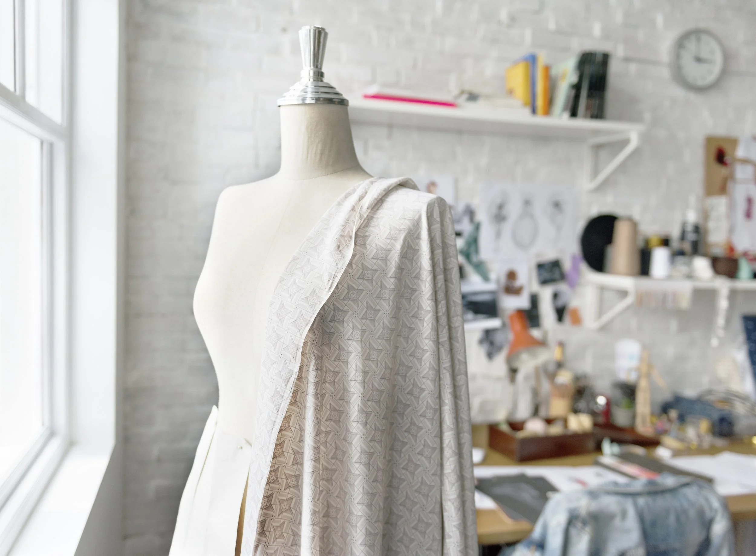 A dress form displaying a piece of fabric in a sewing or design studio with shelves, drawings, and supplies in the background.