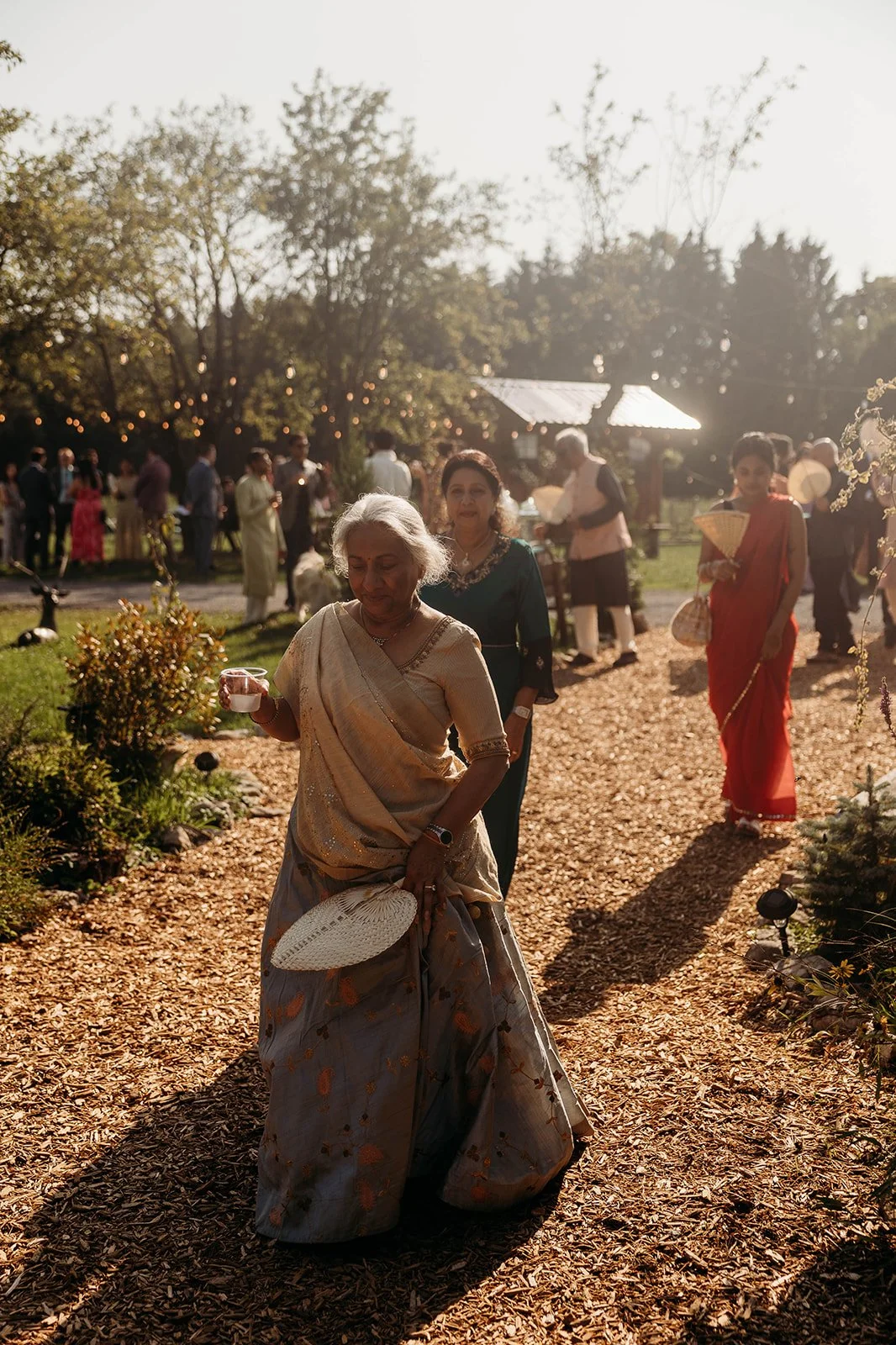 A group of diverse women in traditional Indian sarees walking along a garden path at sunset during an outdoor celebration.