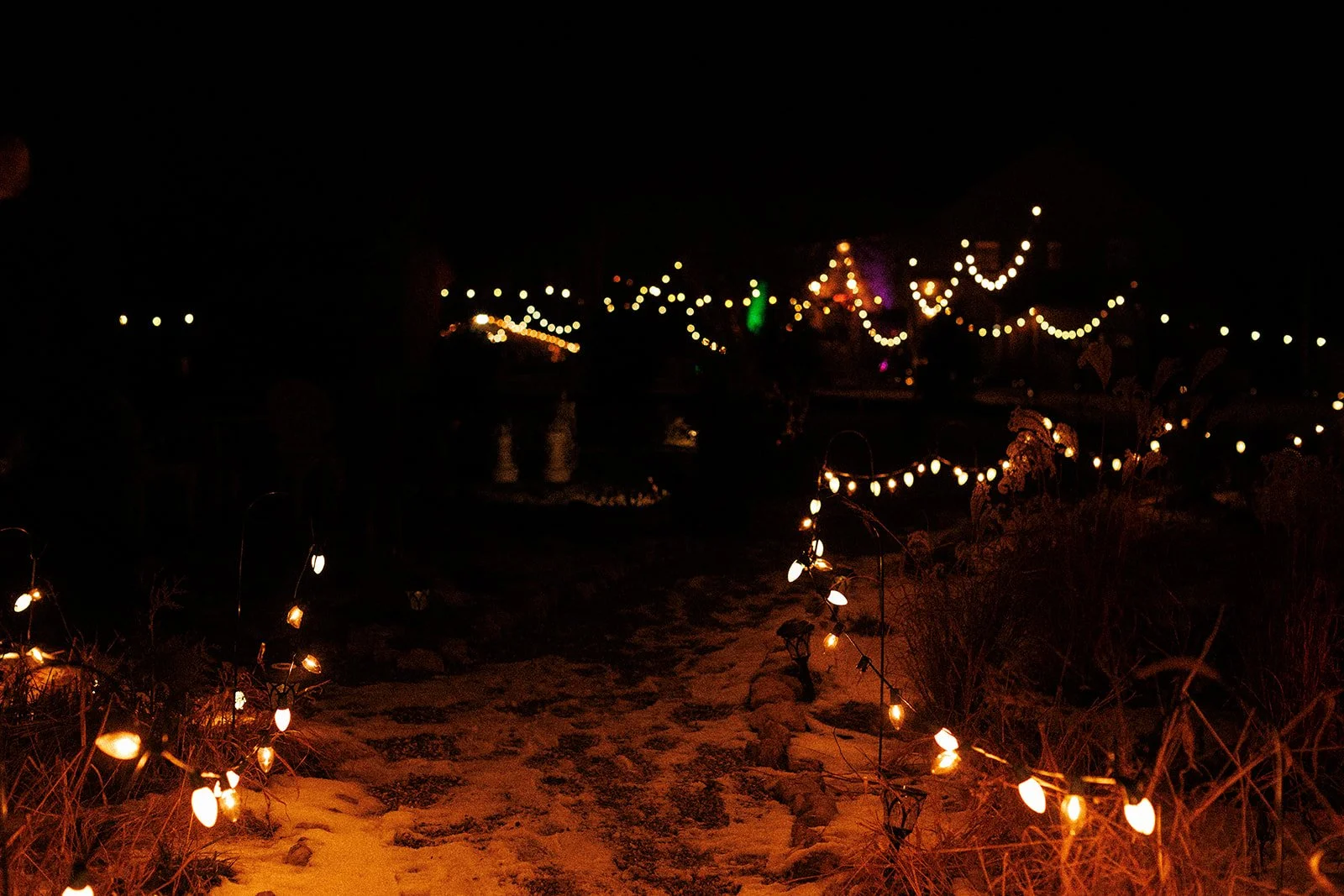 Night scene of a pathway lit with string lights, with additional decorative lighting in the background, creating a festive or romantic atmosphere.