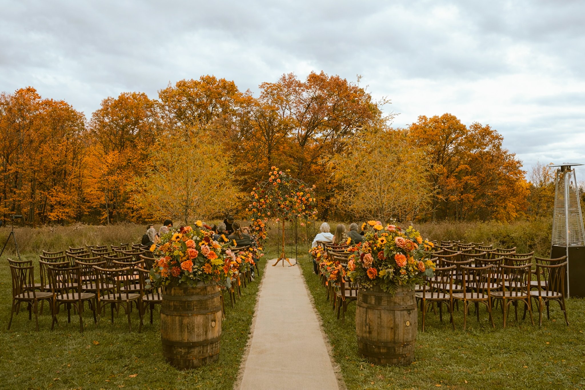 Outdoor wedding ceremony setup with rows of chairs, large floral arrangements in barrels, and a flower arch on a grassy field with trees in autumn colors under a cloudy sky.