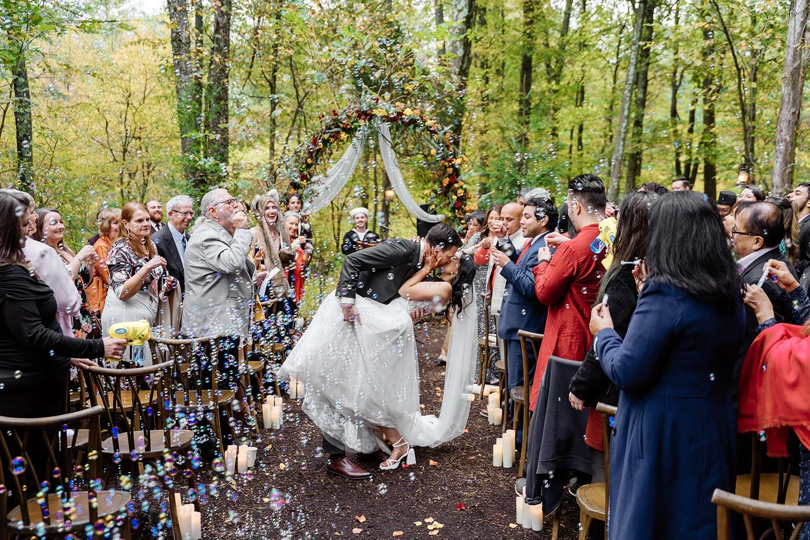A wedding ceremony taking place outdoors in a forest, with a bride and groom kissing as bubbles are blown around them. Guests are smiling and cheering, and there is a floral arch in the background.