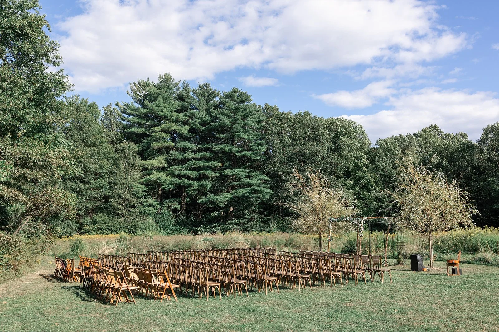 Outdoor wedding ceremony setup with rows of wooden chairs facing a simple arch with trees and greenery in the background, under a partly cloudy sky.