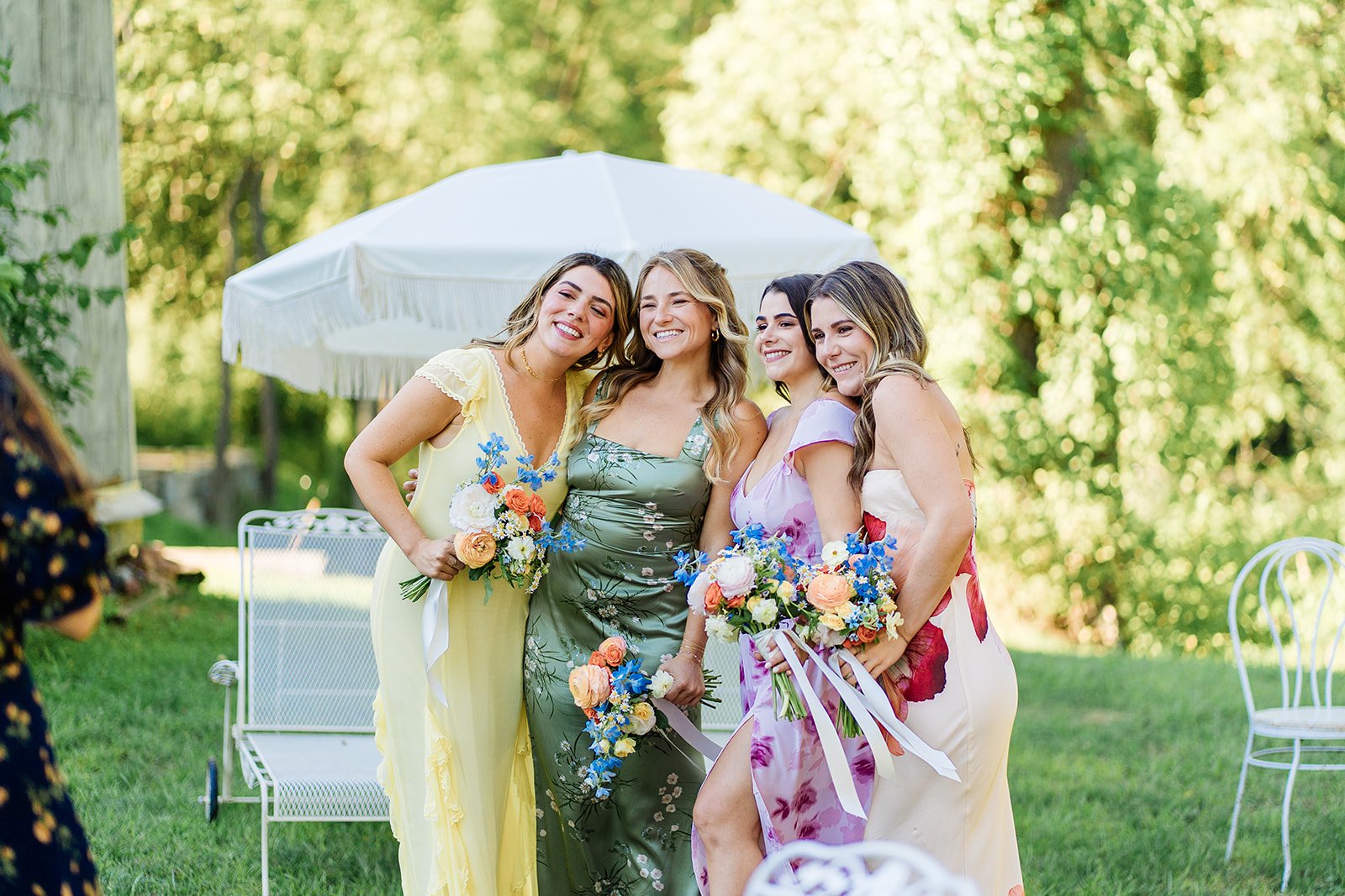Four women in colorful dresses holding bouquets, smiling and posing outdoors under a white umbrella, with green trees and grass background.
