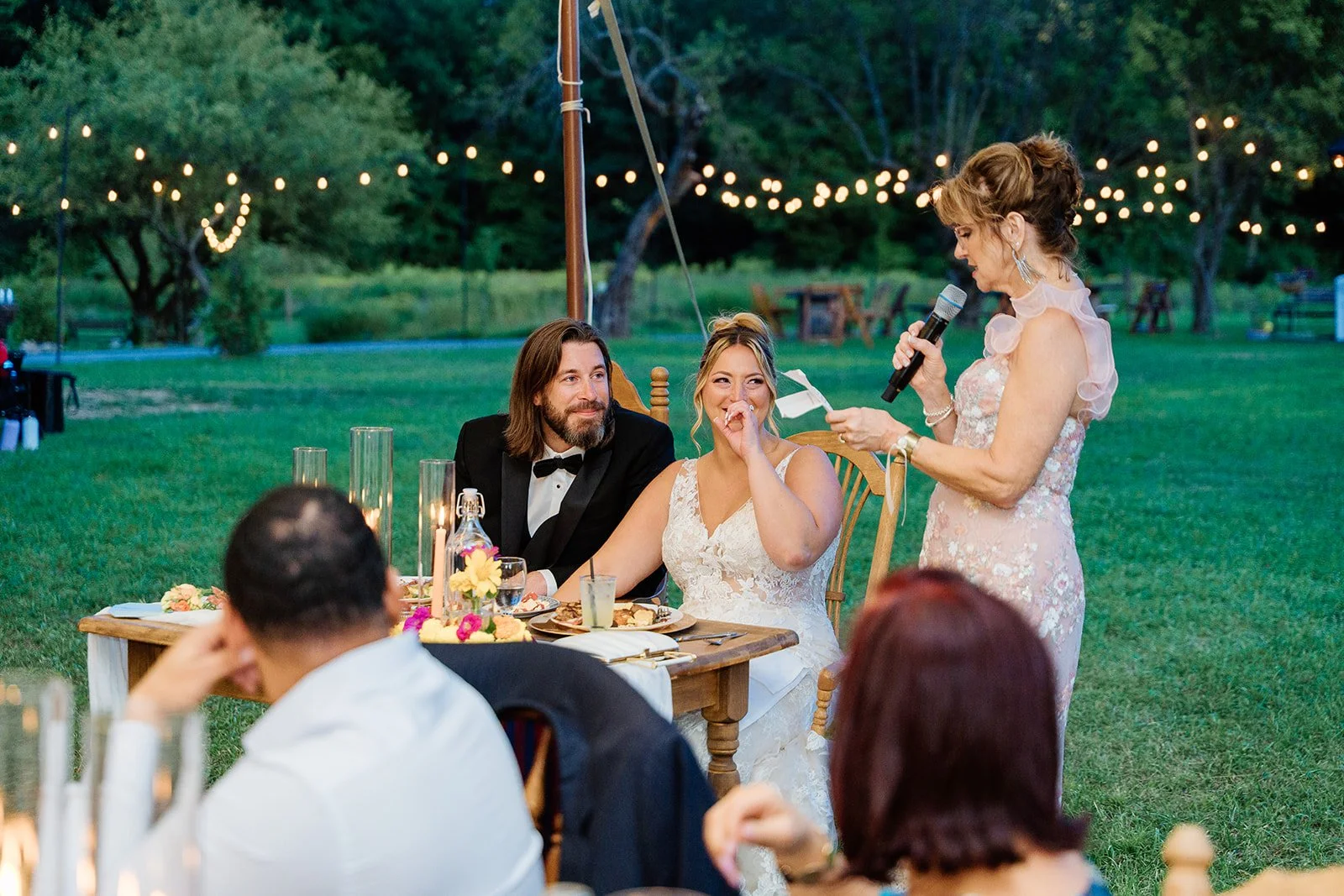A wedding reception outdoors at dusk with string lights overhead. A woman in a light pink dress is giving a speech into a microphone while a bride and groom look on. The bride is laughing and covering her mouth with her hand, wearing a white lace gow