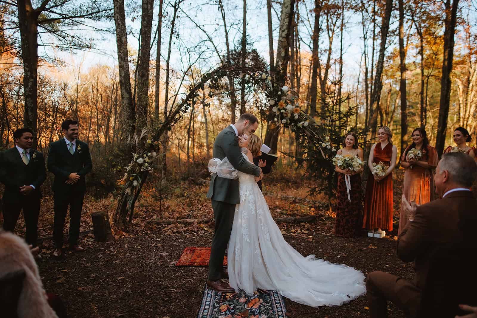 A wedding ceremony taking place outdoors in a forest at sunset. A couple is hugging under a decorated floral arch, surrounded by their wedding party with bridesmaids holding bouquets and groomsmen dressed in suits.