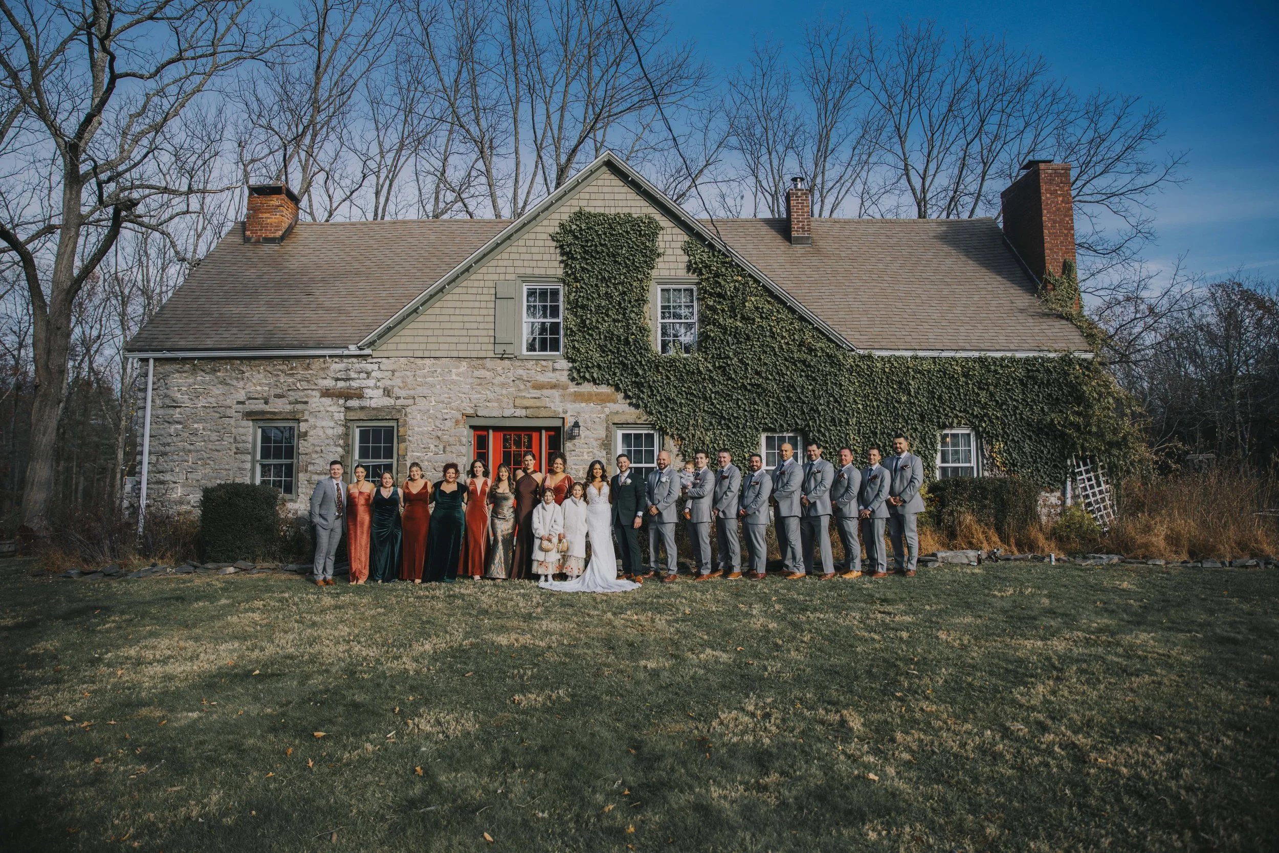 A large group of people dressed in formal attire standing outside in front of an old stone and wood house with vines growing on it, during daytime with leafless trees in the background.