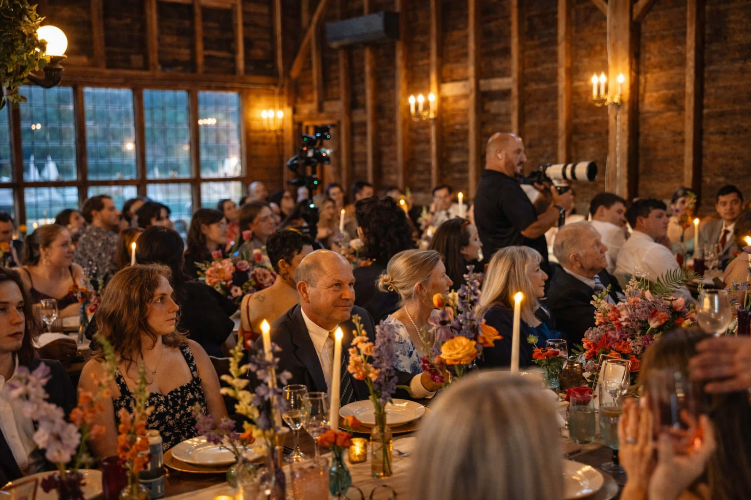 Guests seated at a decorated banquet table in a rustic wooden hall, listening to a speaker or performer with a camera, with floral arrangements and lit candles on the table.