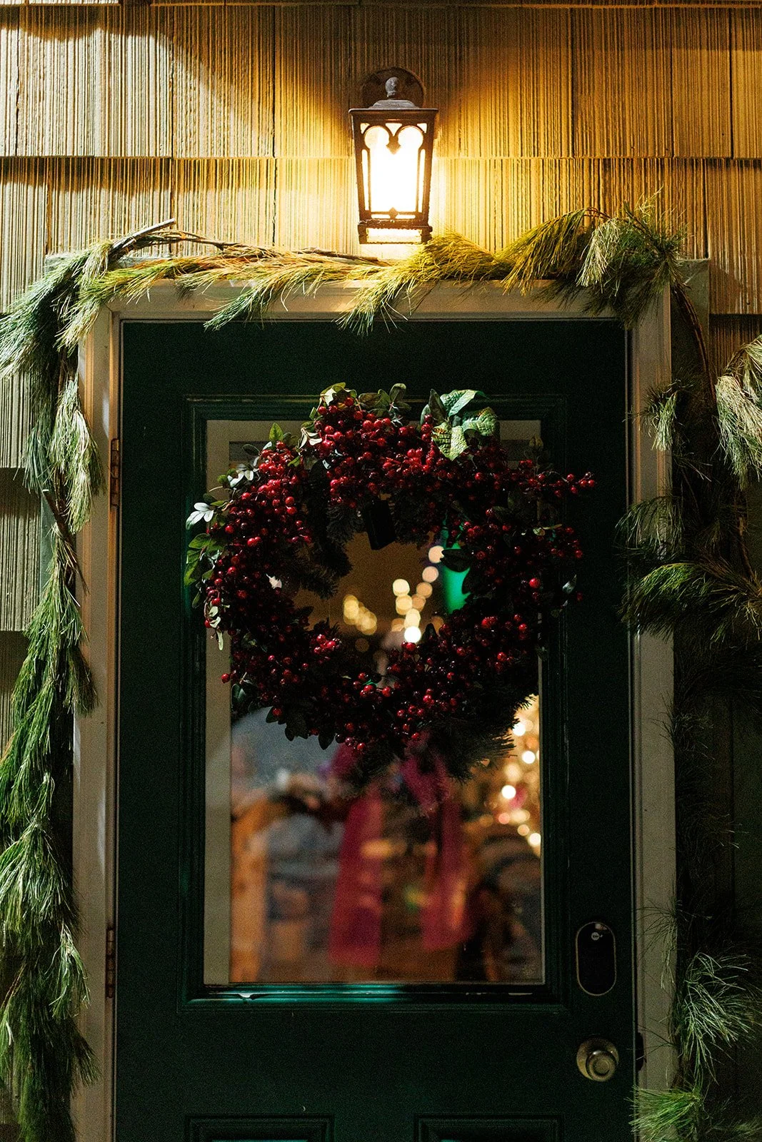 A Christmas wreath decorated with red berries hanging on a green door, with a Christmas garland and a lantern above it. Warm indoor lights are visible through the glass door.