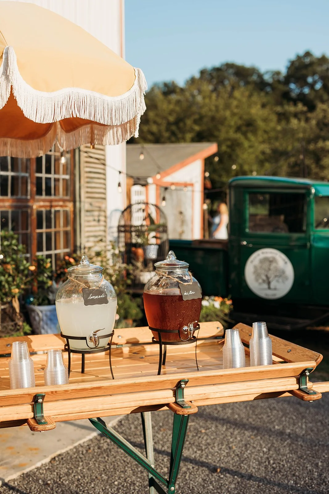 Outdoor beverage stand with glass dispensers of lemonade and iced tea, and clear plastic cups, shaded by a large beige umbrella, with a green truck and a rustic building in the background.