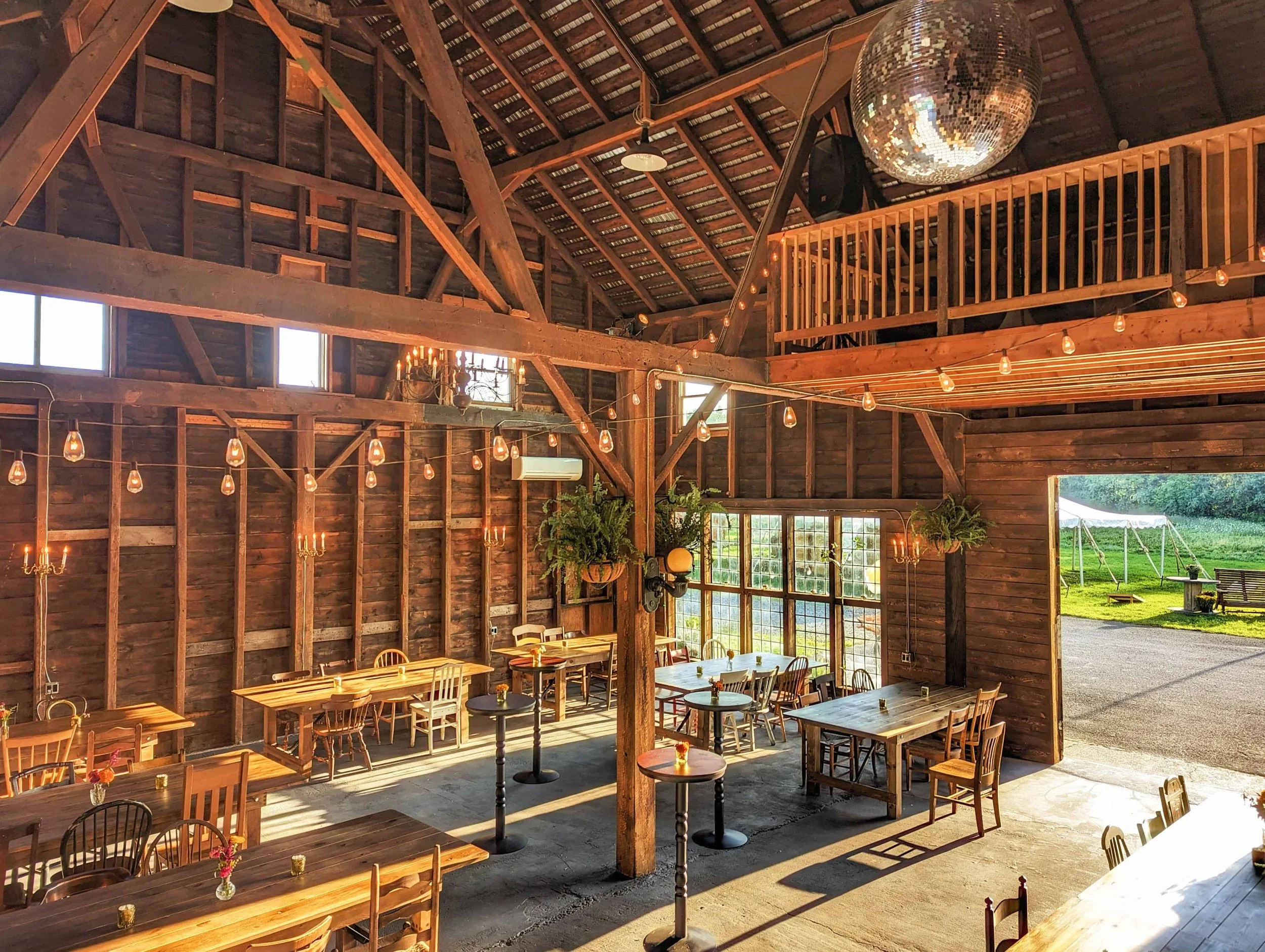 Interior of a rustic barn converted into a dining area with wooden tables and chairs, string lights, potted plants, large windows, and an open door leading outside to a grassy area with a swing and a bench.