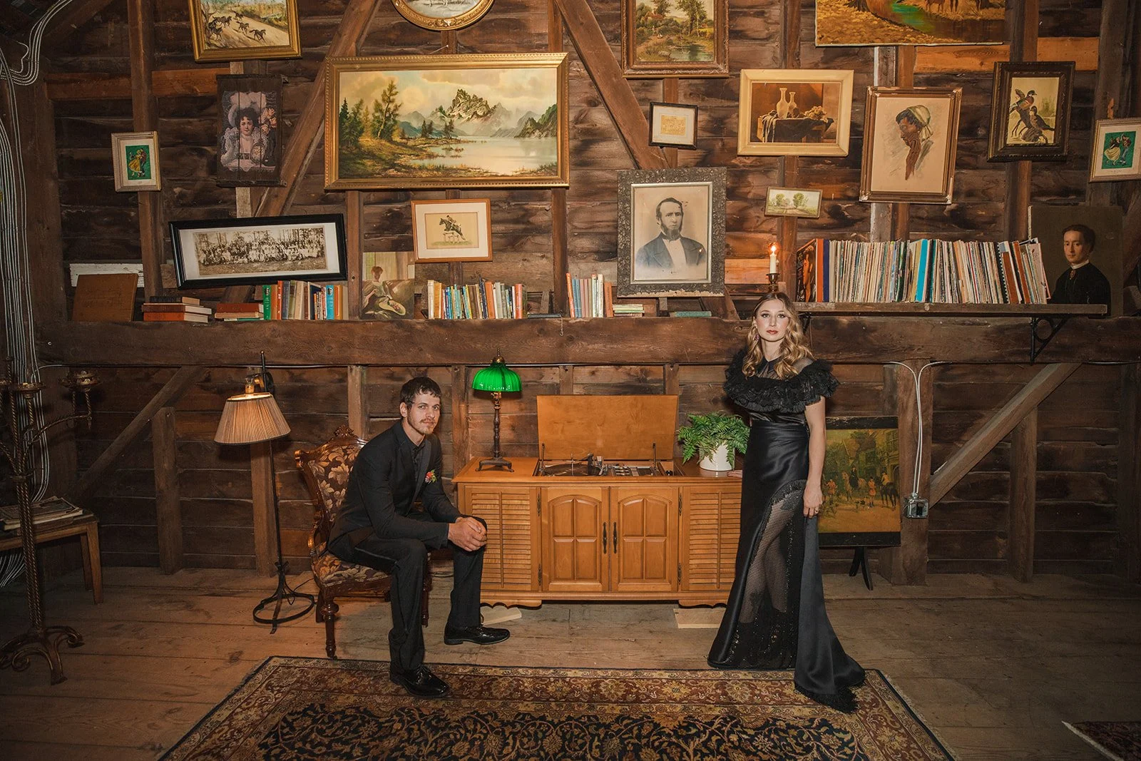 A man and a woman in vintage-style clothing pose inside a rustic wooden room decorated with numerous framed paintings, books, and antique furniture. The man sits on a floral upholstered chair near a small wooden cabinet with a green-shaded lamp, whil