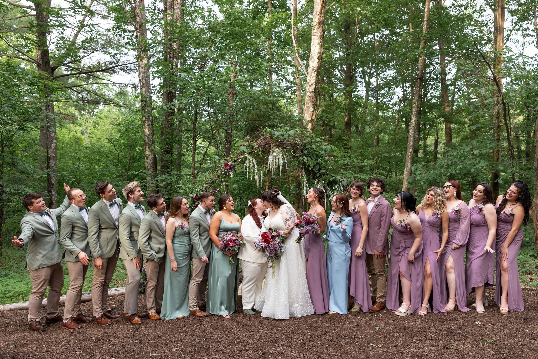 A wedding party stands in a forest, with the bride and groom at the center, surrounded by bridesmaids and groomsmen all dressed in pastel shades, celebrating during daytime.