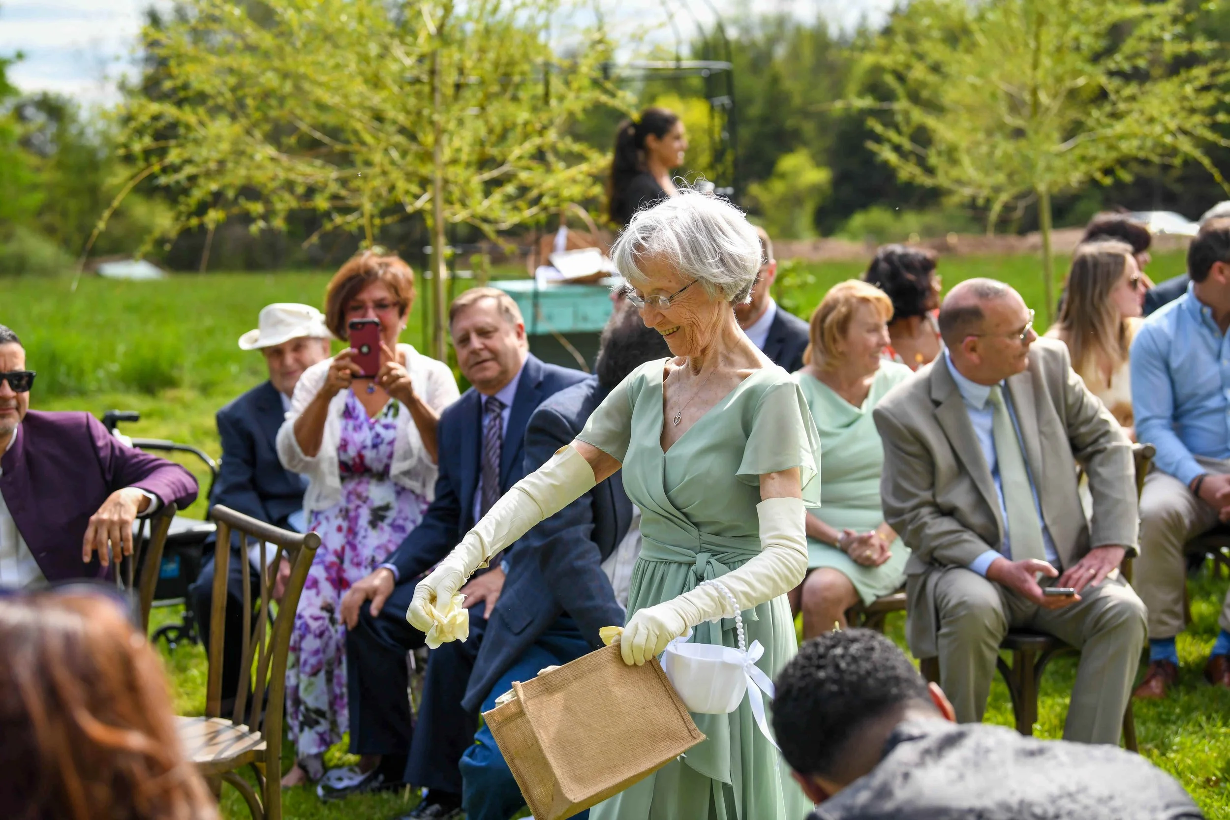 An elderly woman in a mint green dress and gloves smiling as she walks with a basket, at an outdoor wedding ceremony surrounded by seated guests in a grassy park with trees.