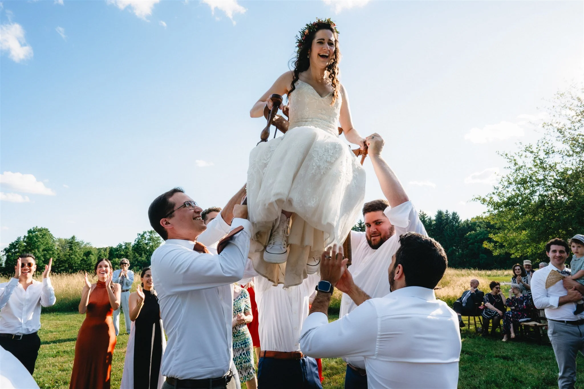 Bride being lifted on a chair during a wedding celebration outdoors with guests clapping and cheering.