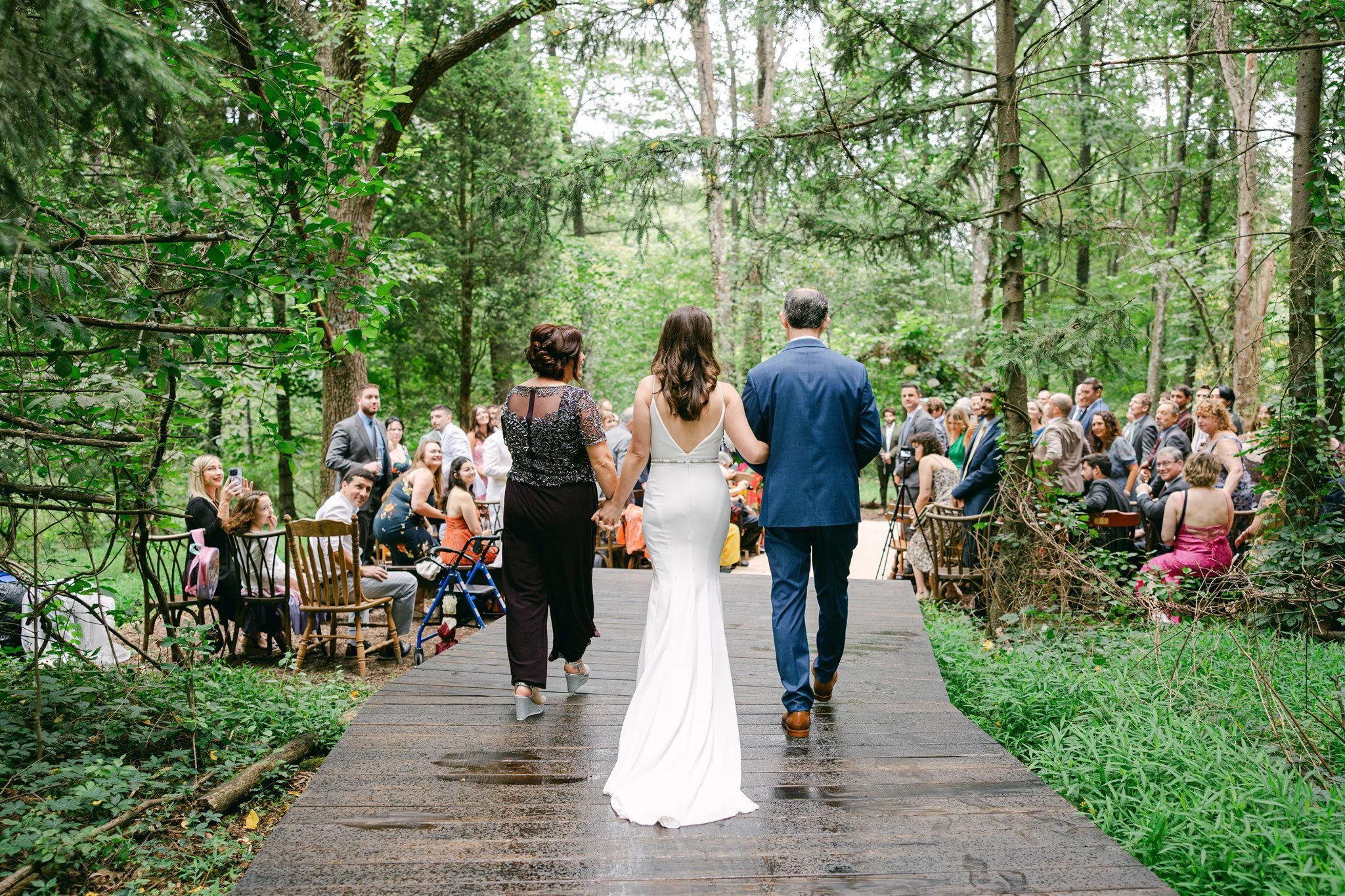 Bride and her parents walking down the aisle at an outdoor wedding ceremony in a lush green forest, with guests sitting and standing on either side, watching and taking photos.