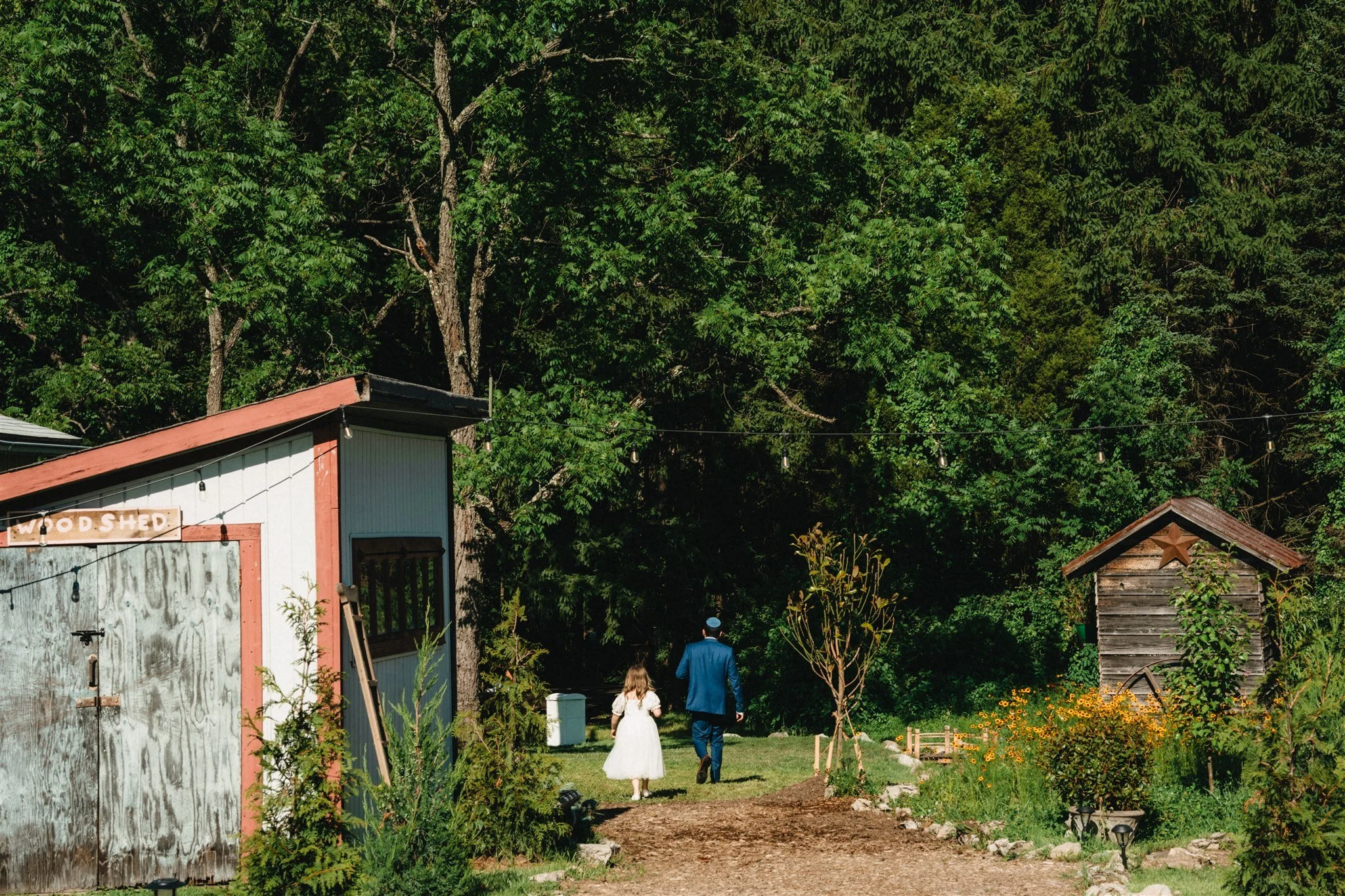 A man in a blue suit and kippah walking with a young girl in a white dress along a dirt path in a garden. In the background, there are green trees, a small wooden shed with a star decoration, and flowers, under string lights.