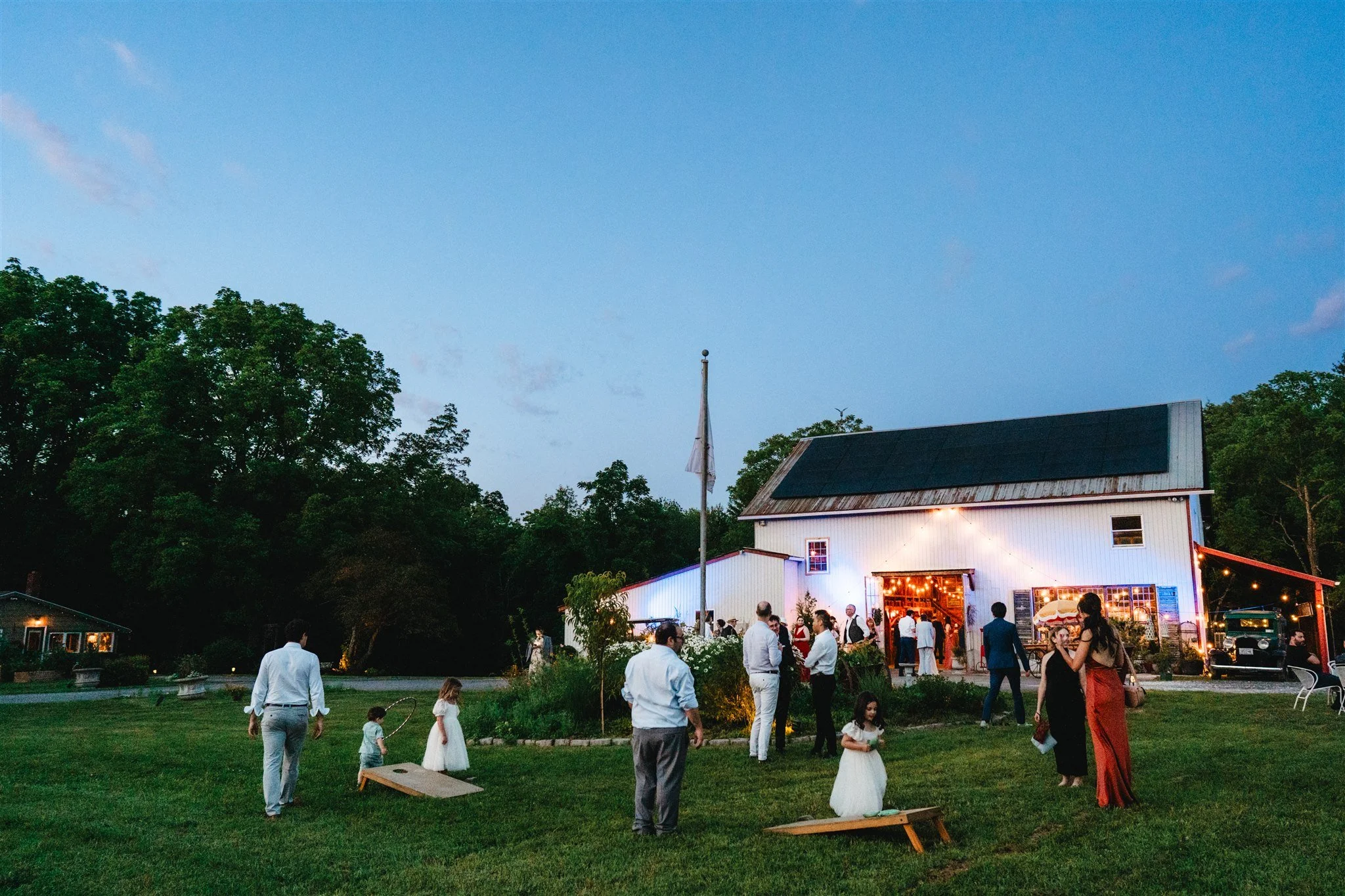 Outdoor gathering with people socializing on a grassy lawn near a white barn decorated with string lights in the evening sky, surrounded by trees.