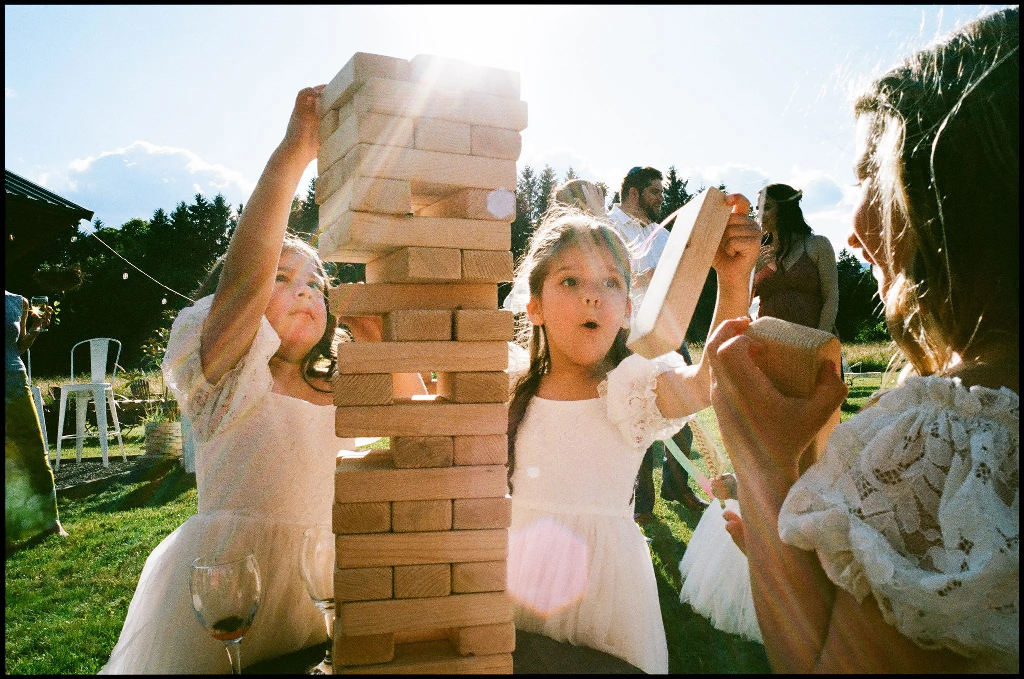 Children at an outdoor wedding or party playing a giant Jenga game with a large wooden block tower, with adults and trees in the background on a sunny day.
