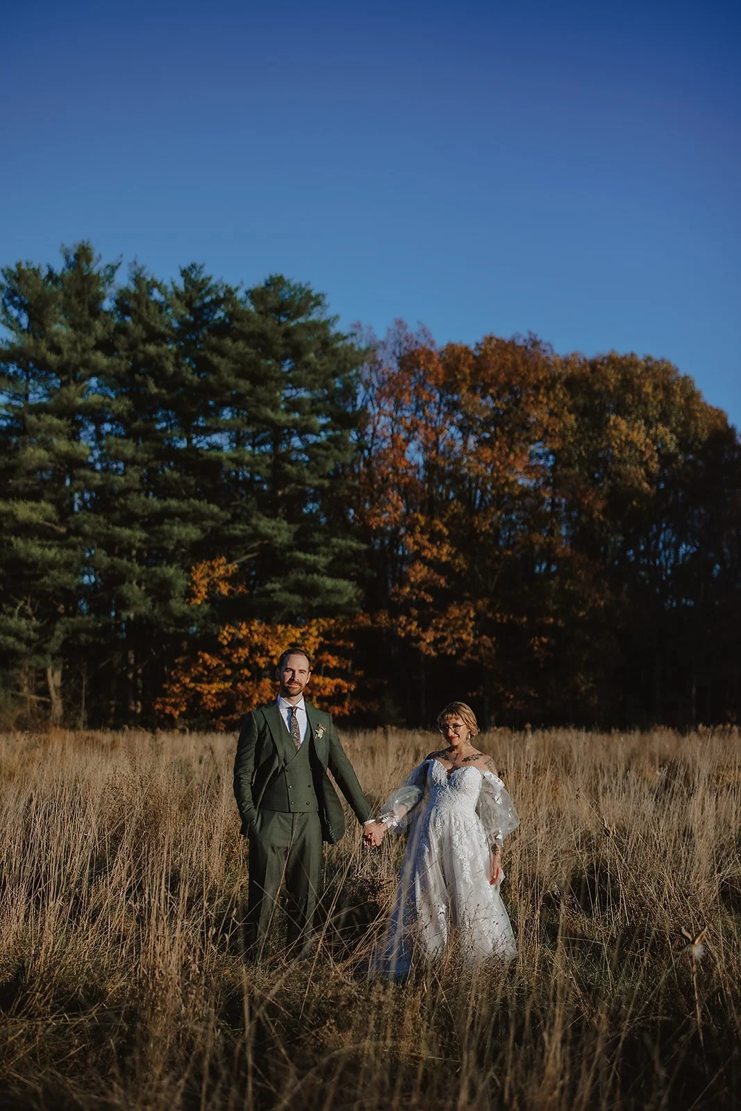 A bride and groom standing hand in hand in a grassy field during autumn with trees in the background.
