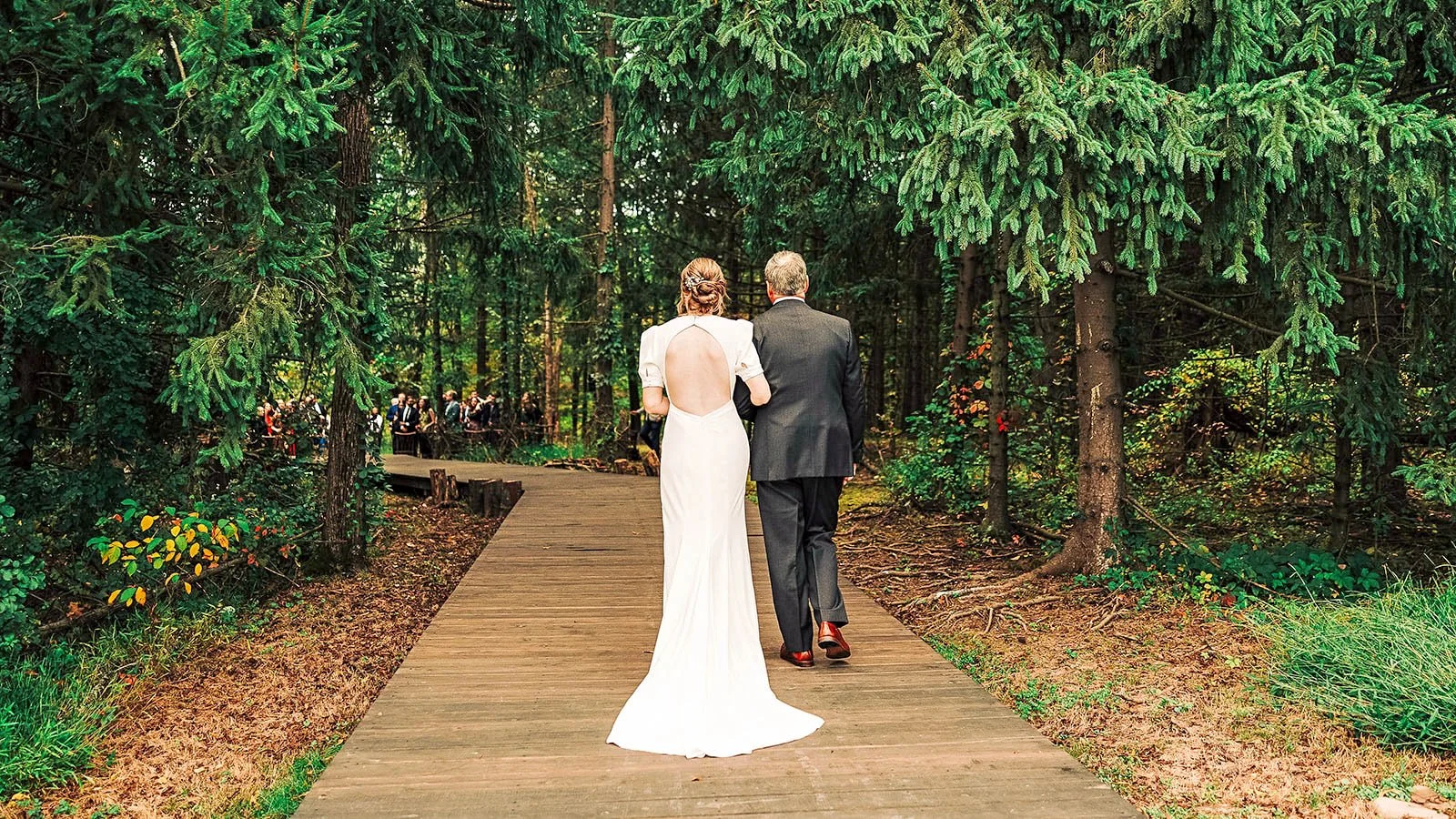 A bride and an older man walking on a wooden path through a dense green forest with wedding guests visible in the background.