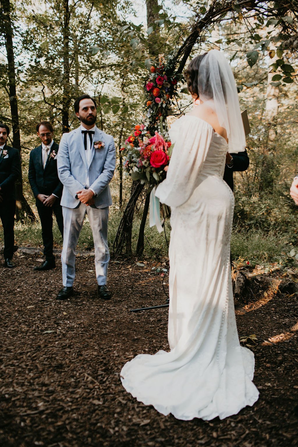 A bride and groom exchange vows during an outdoor wedding ceremony in a wooded area, with a floral arch and three groomsmen in the background.