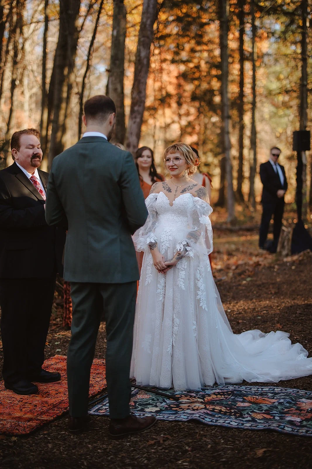 A couple exchange vows during an outdoor wedding ceremony in a forest during autumn. The bride wears a white lace gown with off-the-shoulder sleeves and looks happily at the groom, who is dressed in a gray suit. An officiant stands nearby, and weddin