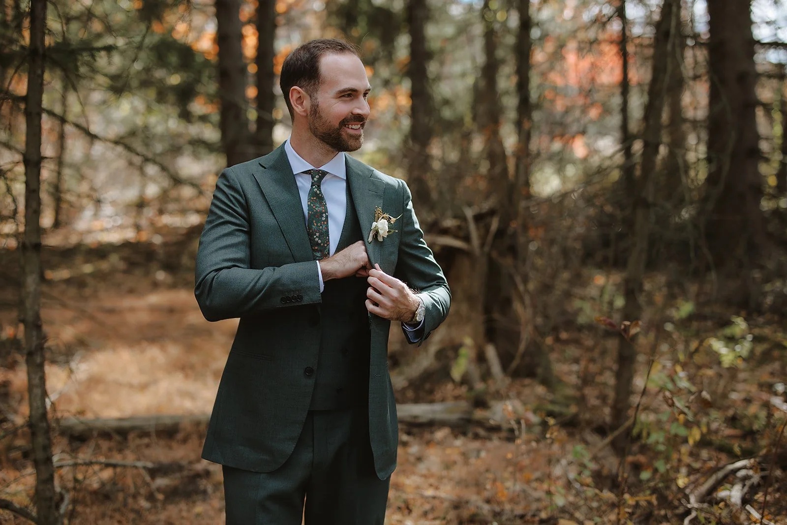 A man in a green suit adjusting his jacket in a wooded outdoor setting, smiling and looking to the side.