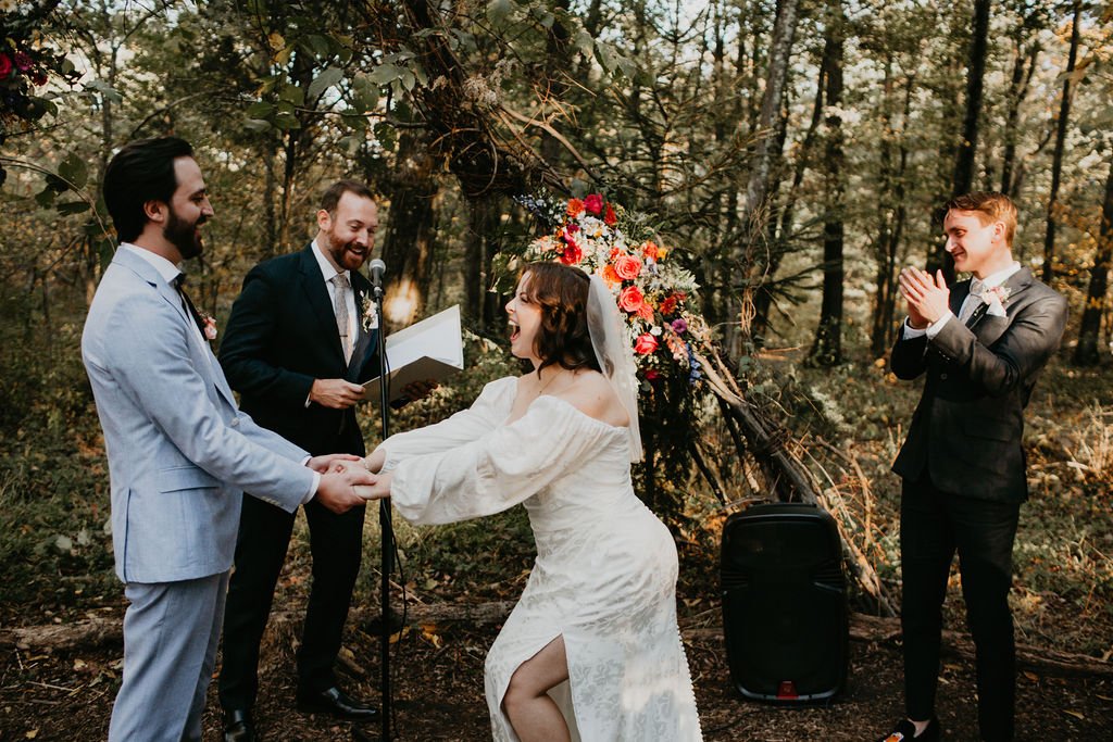 A couple gets married in a forest, holding hands and smiling at each other. The officiant stands behind them with a microphone, while another man claps and smiles to the side. Decorated flowers are visible in the background.
