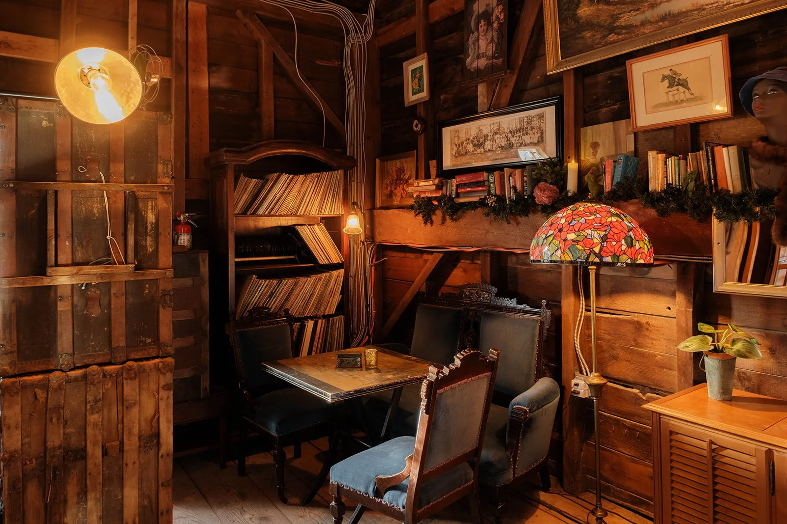 Cozy wooden room with bookshelves, framed pictures, and a table with chairs. Warm lighting from a floor lamp and wall sconce adds to the inviting atmosphere.