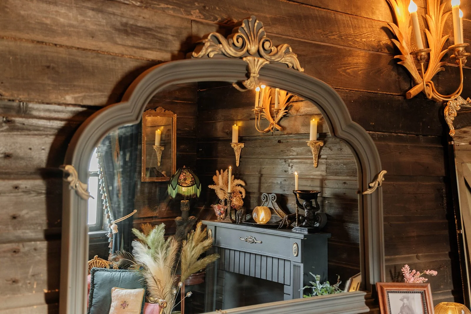 Reflection of a decorated vintage interior in a mirror showing candles, a lamp, framed picture, and various decorative items on a fireplace mantel against a dark wooden wall.